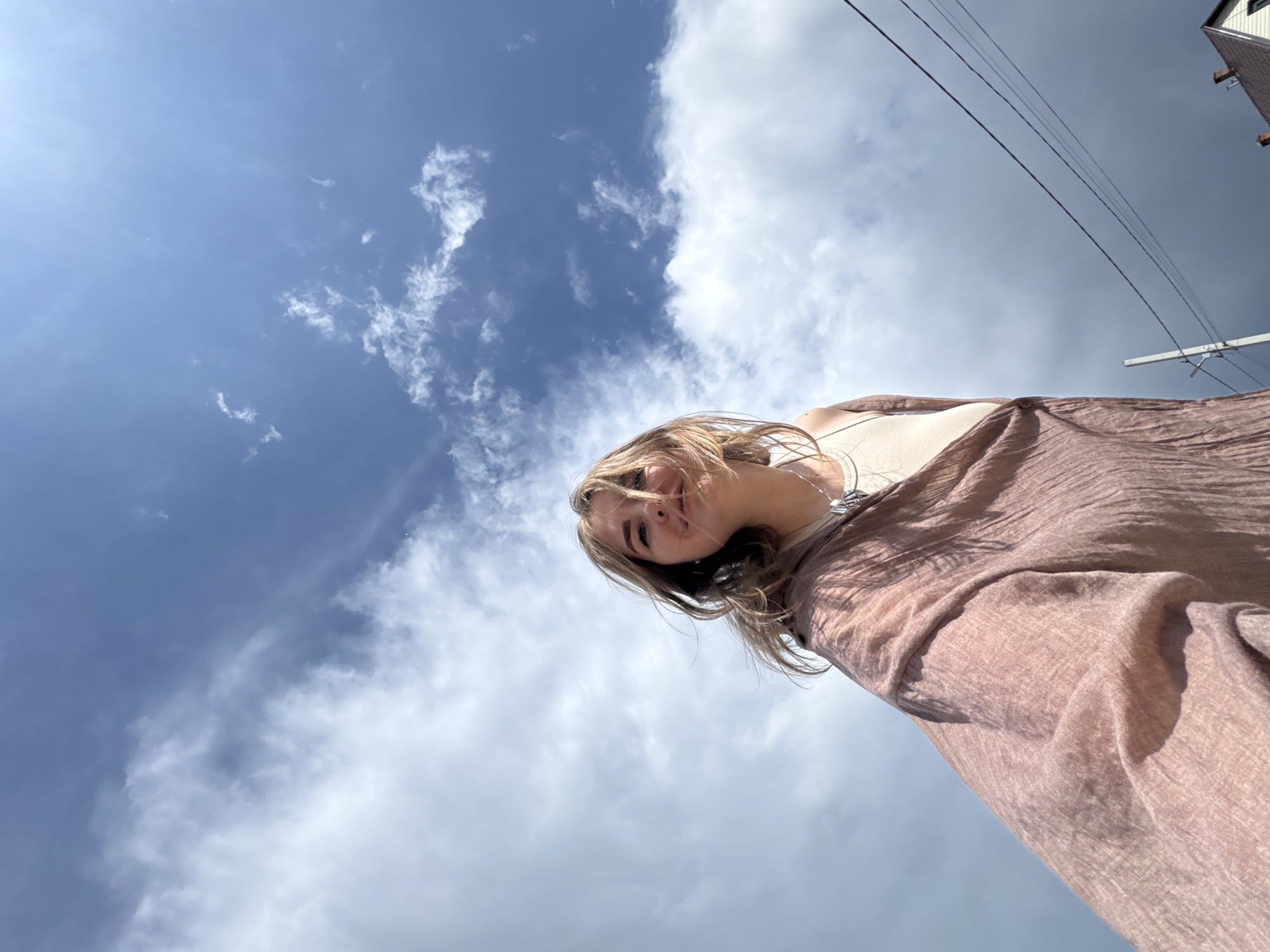 A low-angle, upward-facing photograph depicts a woman against a sky of blue and white clouds. The woman is positioned in the upper portion of the frame, looking down at the camera. She has light hair and is wearing a light-colored top under a brown, textured shawl or scarf. A necklace is visible around her neck. Above and to the right of the woman, there are overhead power lines against a backdrop of partly cloudy sky. A small section of a building with a gable roof is also visible in the upper right corner. The photograph appears to have been taken outdoors during daylight hours in Горный, Russia.