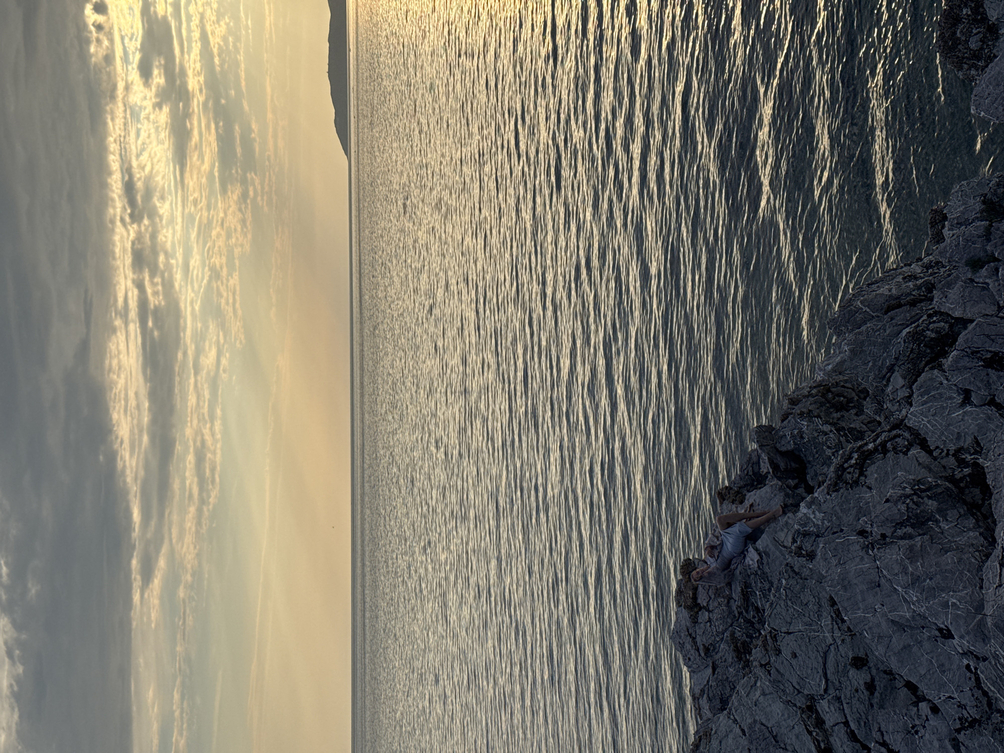 A person is lying on a rocky cliff overlooking the sea at sunset. The sun is low in the sky, casting a golden light on the water and clouds. The sea is calm, with gentle ripples reflecting the light. The rocky cliff is dark and rugged, with sharp edges and crevices. The person is wearing light-colored shorts and appears relaxed, with their legs bent and arms resting on the rocks. The scene is serene and peaceful, with a sense of solitude and contemplation. The location appears to be coastal, with a mountainous coastline visible in the distance on the left side of the frame. The time of day is sunset, indicated by the warm, golden hues in the sky and on the water. The weather is clear, with some scattered clouds that are illuminated by the setting sun. There is no visible text in the image.