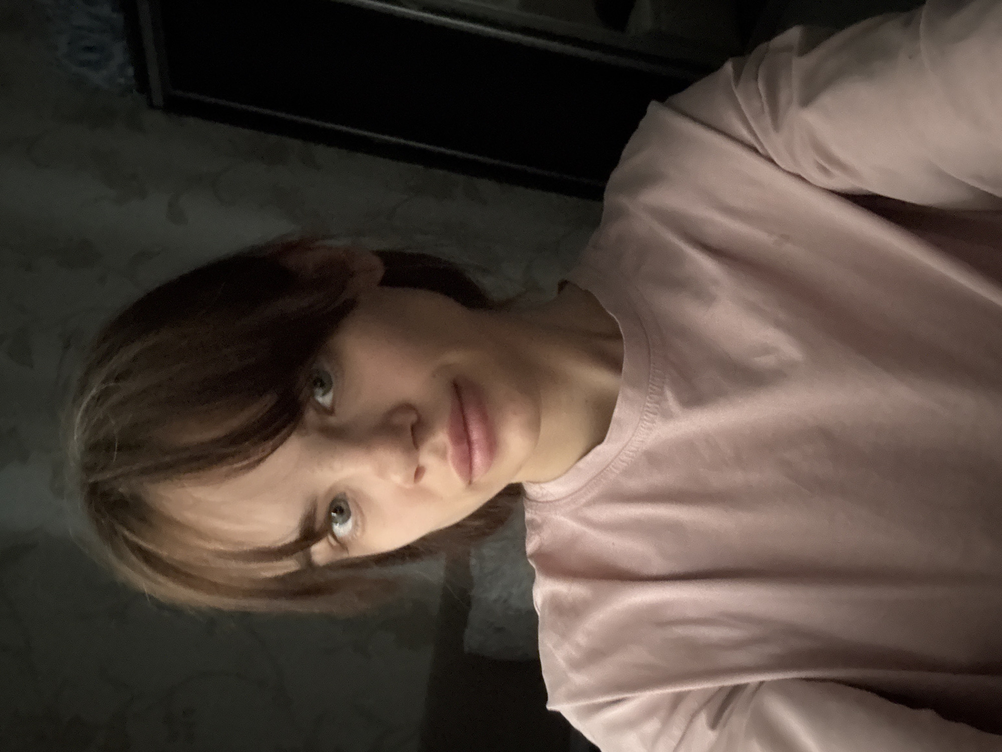 A selfie-style portrait shows a young person in an indoor setting, rotated 90 degrees clockwise. The subject, likely female, has light to medium brown hair with lighter highlights, styled with bangs that partially cover her forehead. Her eyes are light-colored (appearing blue or green) and are directed towards the camera. She has a neutral to slightly pensive expression, with her lips slightly parted.
She is wearing a plain, light pink, long-sleeved t-shirt or top. The fabric shows some wrinkles, particularly around the shoulder and arm.
The background is dimly lit and somewhat indistinct. To the upper left and behind the subject's head, there's a subtly patterned wallpaper. A dark, rectangular object, possibly furniture, a mirror frame, or a window frame, is visible behind and above the subject's right shoulder. The overall lighting suggests an indoor environment, possibly in the evening or with artificial light. There are no direct visual cues to the specific location of Rostov-on-Don, Russia, within the image. No text is visible.