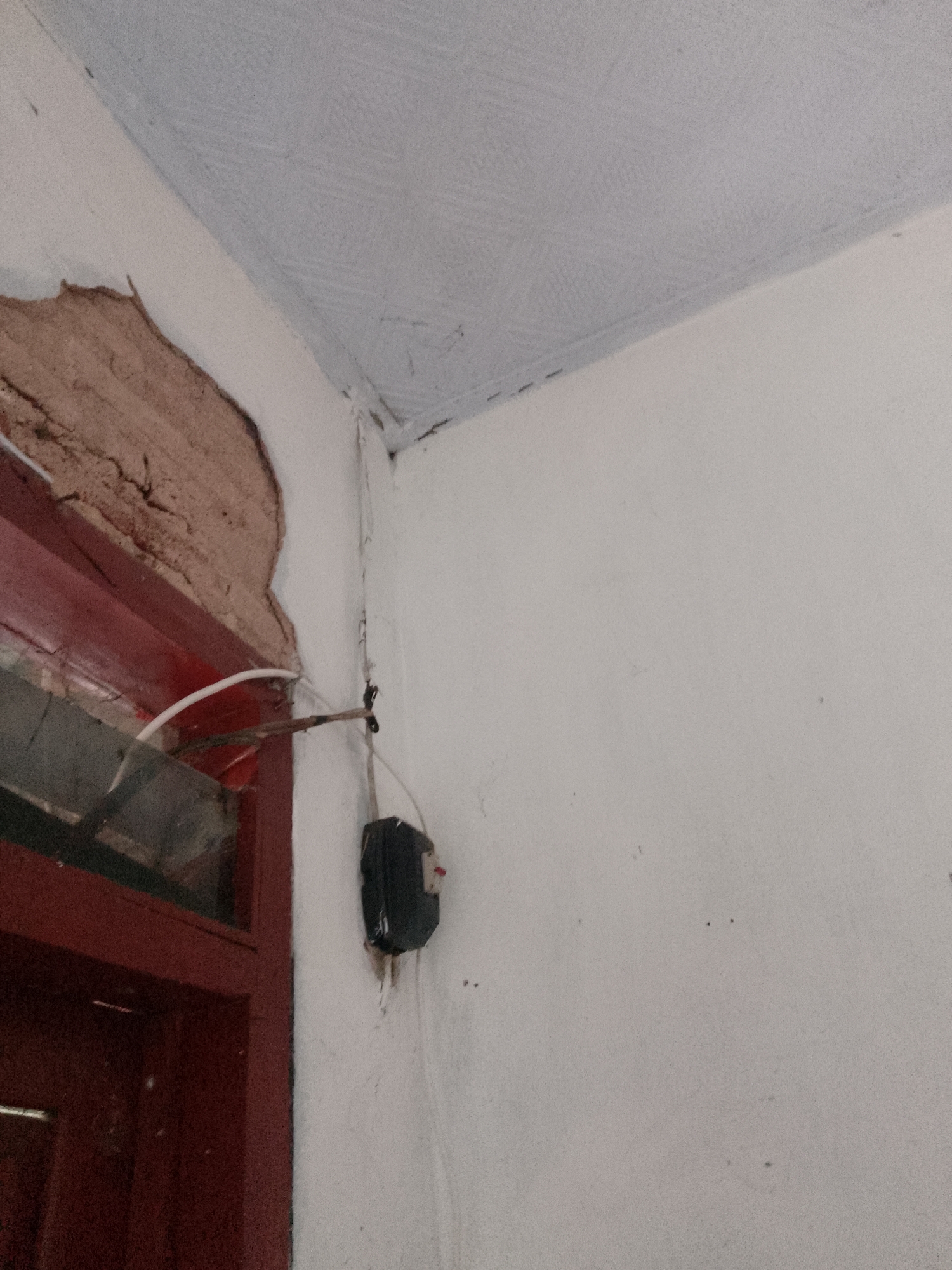A close-up, eye-level shot captures a corner where a white wall meets a pale blue, textured ceiling. The plaster on the wall is damaged and peeling away, revealing a rough, reddish-brown interior layer. A dark red door frame is partially visible on the left, with a section of a dark paneled door beneath it. A metal rod protrudes horizontally from the wall near the damaged plaster, with a white tube attached to it. Further down the wall, a black electrical box is mounted, featuring a red switch. White electrical wires run along the wall and ceiling. The lighting is dim, suggesting an indoor environment. The image does not contain any identifiable landmarks or context that would allow for a specific location within Trenggalek, Indonesia.