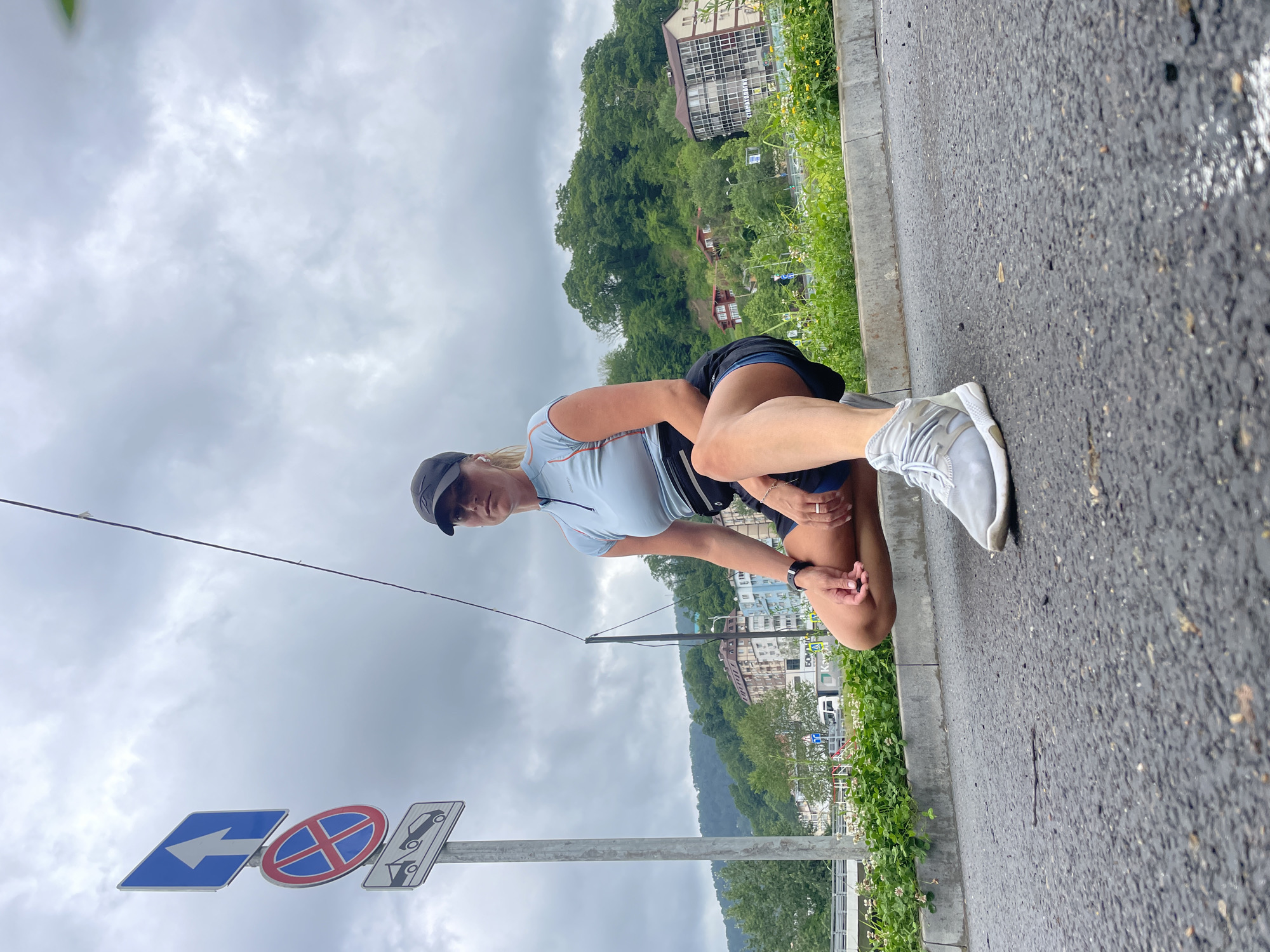 A female is kneeling on the asphalt edge of a road in Sochi, Russia. She is wearing a light blue, short-sleeved athletic shirt, dark shorts, a black cap, and white athletic shoes. She is looking at the camera while seemingly stretching her leg. Road signs are visible on the left side of the image, including a blue directional arrow, a red "no stopping" sign, and a parking assistance sign. A concrete sidewalk edge borders the road, with grass and foliage growing beside it. Buildings and green forested hills are in the background, under a cloudy sky.