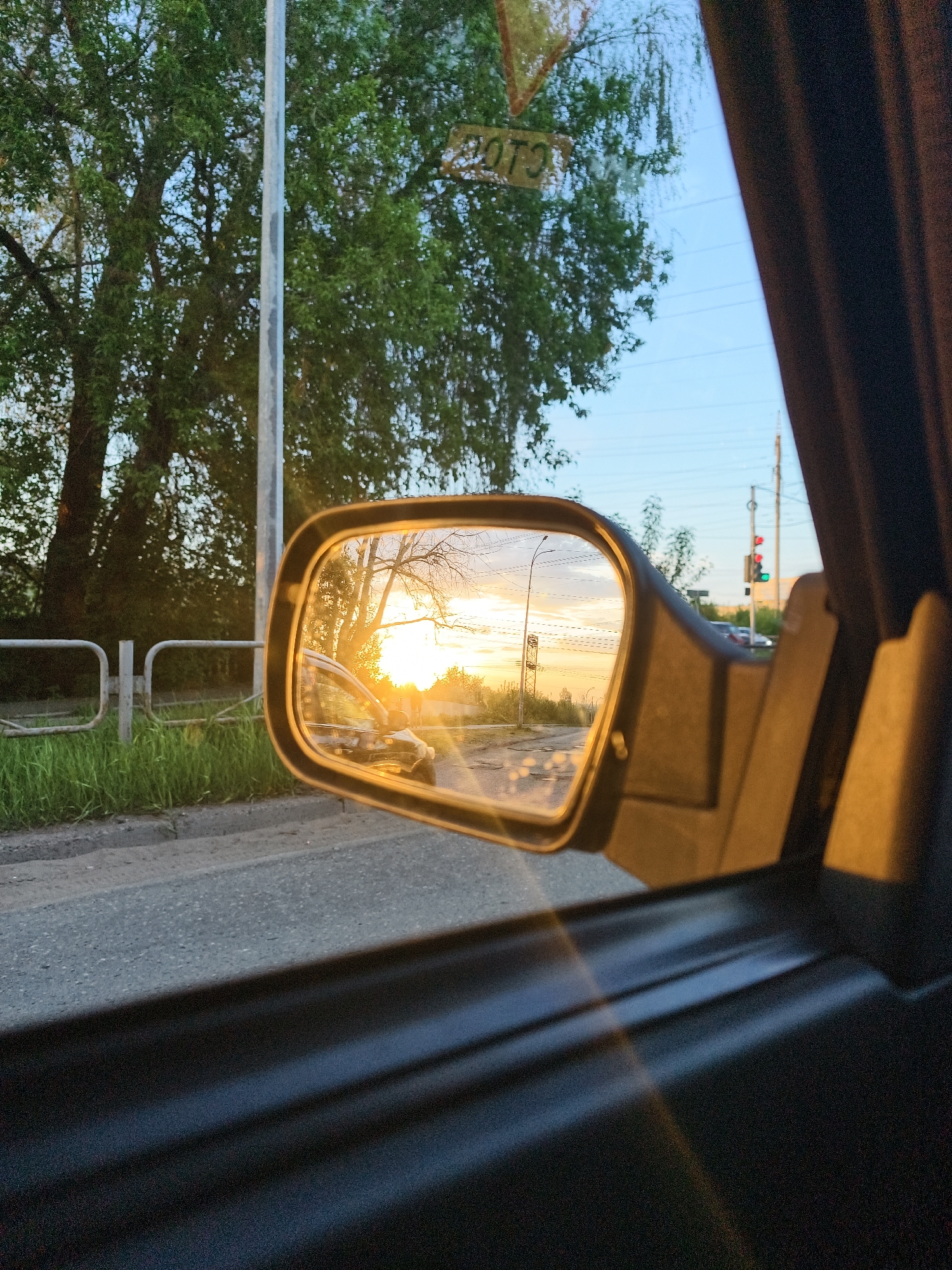 A view from inside a vehicle looking out at a sunset mirrored in a side mirror. The exterior view shows a street with a sidewalk, a metal railing, grass, and trees. The side mirror reflects a setting sun over a road. A street light and traffic lights are visible further down the road. The reflection includes another vehicle behind the car taking the photo. The image appears to be taken in or near Chaikovsky, Russia, given the presence of Cyrillic script on a sign reflected in the window.