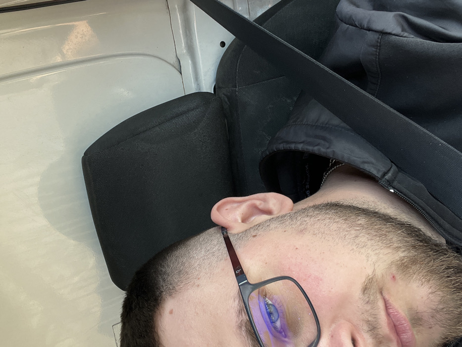 A male individual is depicted in a close-up, angled portrait within a vehicle in Koilovtsi, Bulgaria. The subject, visible from the forehead to the upper chest, has short, dark hair with a faded cut, light stubble, and blue eyes. He wears black-framed rectangular glasses, with a distinct purple-blue reflection on the left lens. A small mole is present above his left eyebrow, and a red blemish is visible on his chin. He is dressed in a black zip-up jacket, under which a silver-colored chain is visible around his neck. A black seatbelt extends diagonally across his chest and shoulder. Behind him is a black fabric headrest and seat back, consistent with a vehicle interior. The background features a light-colored, utilitarian paneling, suggesting the inside of a van or similar vehicle, with subtle textures and reflections from ambient lighting.