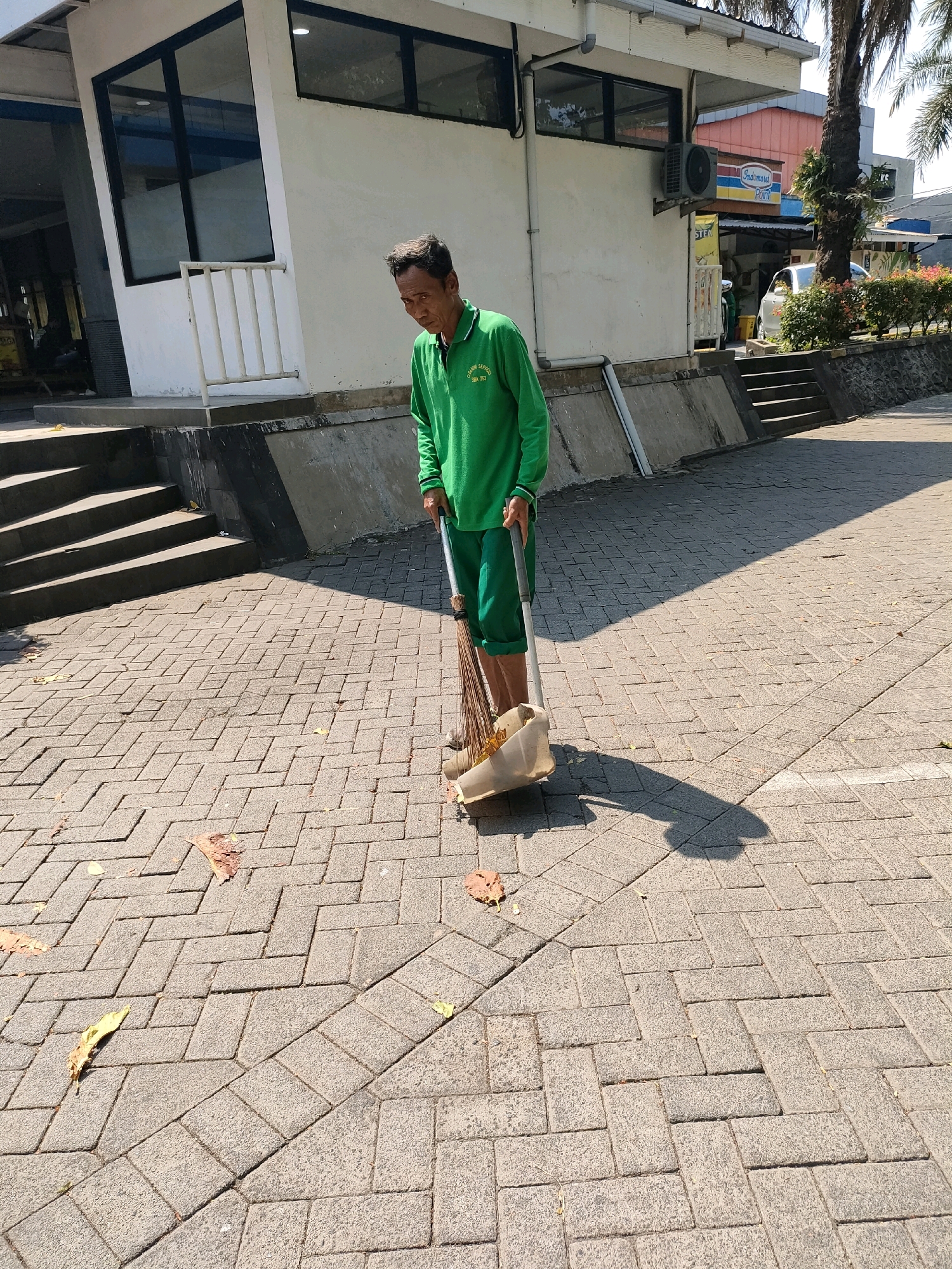 A male sanitation worker in Sukodono, Indonesia, wearing a green long-sleeved shirt and green pants, sweeps a paved walkway in front of a white building. He holds a broom and dustpan. The building has a modern design with large windows and an air conditioning unit mounted on the exterior wall. A sign for "Indomaret Point" is visible on the building. Palm trees and bushes are present in the background, indicating an outdoor, sunny environment. The ground is covered with interlocking brick pavers, and fallen leaves are scattered across the walkway. Stairs are visible on the left side of the image, leading up to the building's entrance. The overall scene depicts a routine maintenance activity in an urban or commercial setting.