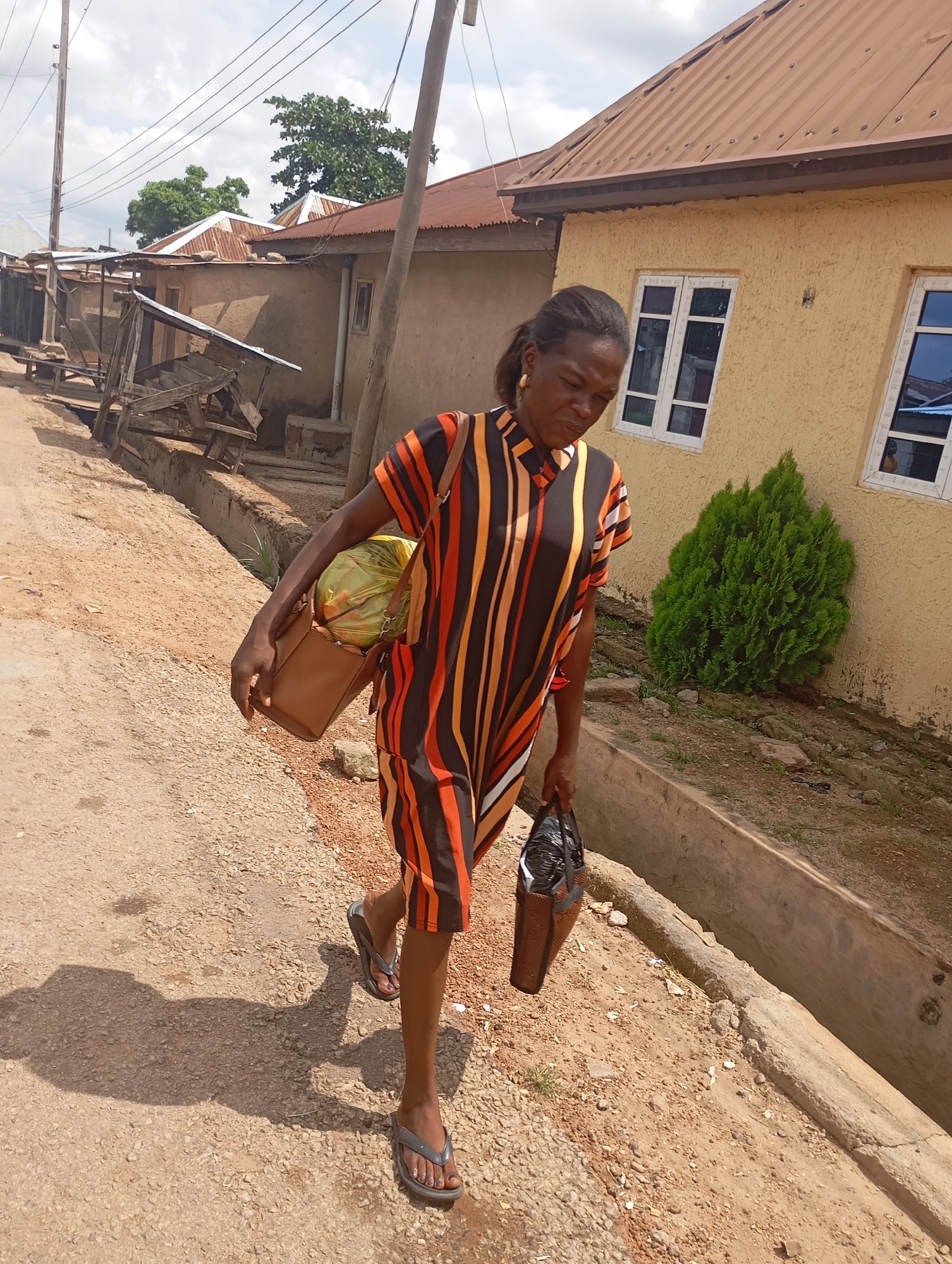 In the city of Shendam, Nigeria, a woman walks along a dirt road, carrying two bags. She is wearing a striped dress with orange, black, and yellow stripes, and appears to be wearing flip-flops. One bag, a brown leather purse, is slung over her left shoulder, and she is carrying a yellow object wrapped in plastic inside it. In her right hand, she holds another brown bag with a black plastic bag inside. The background shows a row of buildings with corrugated metal roofs, and a concrete ditch runs along the side of the road. A small evergreen tree is visible near one of the buildings. The sky is cloudy.