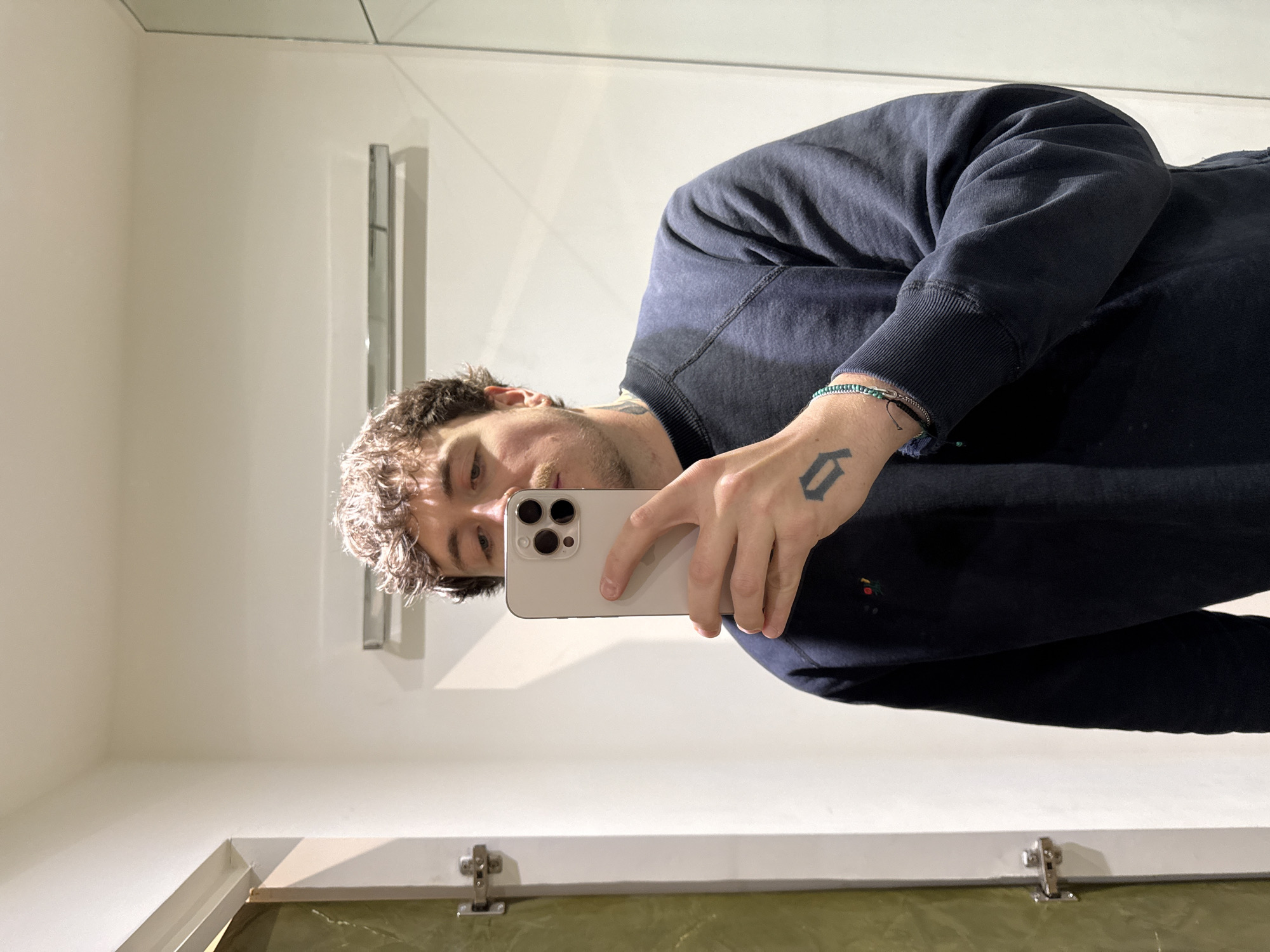 A Caucasian male is taking a selfie in a bathroom mirror. He is wearing a navy-blue sweatshirt and holding a white iPhone. The bathroom has white walls and a mirror. The lower part of the mirror reflects a surface with a slight green hue. The location and city are not discernible.