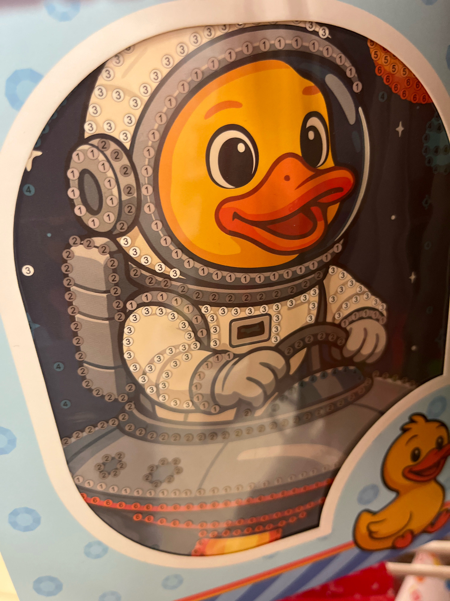 The image is a close-up shot of a paint-by-numbers kit featuring a cartoon duck in a spacesuit. The duck's head is large and yellow with prominent black eyes and an orange beak. It is wearing a white spacesuit with a transparent helmet, revealing its face. The spacesuit is decorated with numbered circles, indicating where specific colors should be applied. The background of the kit is dark blue, speckled with white stars and some blurry circular shapes, suggesting a space theme. On the right side of the main image, a smaller, simpler illustration of a yellow duck is visible. The overall impression is that of a children's craft product.
This image was taken in Kharkiv, Ukraine. The specific location within the city is not identifiable from the image itself.