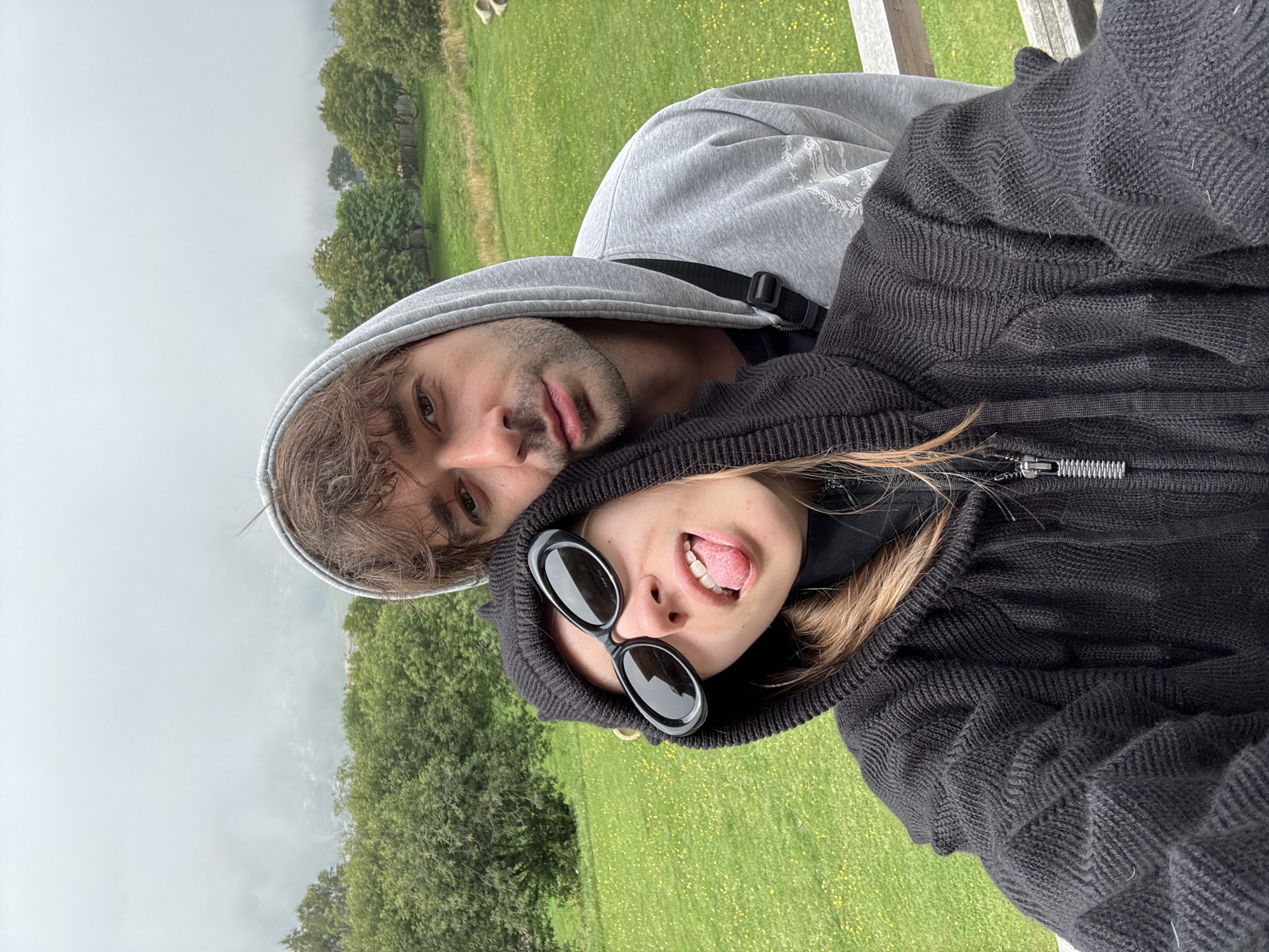 In the city of Stepantsminda, Georgia, a selfie is taken by a young woman and a young man. The woman is in the foreground, wearing a black hooded sweater and black sunglasses, with her tongue sticking out. The man is behind her, wearing a grey hooded sweater and leaning his head against hers. They are outdoors, with a backdrop of green hills, trees, and a foggy sky. A wooden railing is visible in the lower right corner.