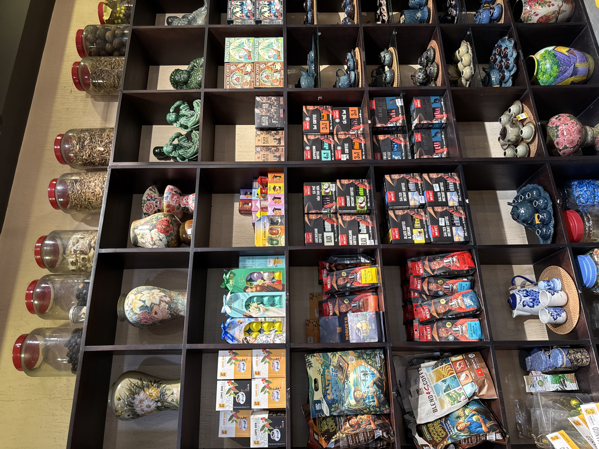 The image shows a densely packed shelf display in what appears to be a shop in Nha Trang, Vietnam. The shelves are filled with various products, primarily coffee, tea, and decorative vases.
In the top section of the shelves, glass jars containing dried ingredients and what appear to be spices or herbs are visible. Below these jars, decorative ceramic vases with floral motifs are displayed. Further down, there are numerous boxes and bags of coffee, with brands like "Cafe Sua," "Mr. Viet," and "Htc" being discernible. Some of these packages feature illustrations of people and coffee beans. Mixed in with the coffee products are some colorful boxes that might contain tea or other beverages.
On the right side of the shelves, there are sections dedicated to decorative items. These include ceramic figurines of what appear to be Buddhist deities or wise men, and several shelves filled with intricate blue and white tea sets, as well as other ceramic teapots and cups. The lower shelves also display larger decorative vases with vibrant, detailed patterns. Some of the coffee bags and boxes are positioned with their labels facing forward, indicating they are for sale.
The lighting suggests an indoor environment, likely a retail space. The overall impression is of a well-stocked shop offering a variety of beverages and decorative items, consistent with a souvenir or specialty food store in a tourist area like Nha Trang.