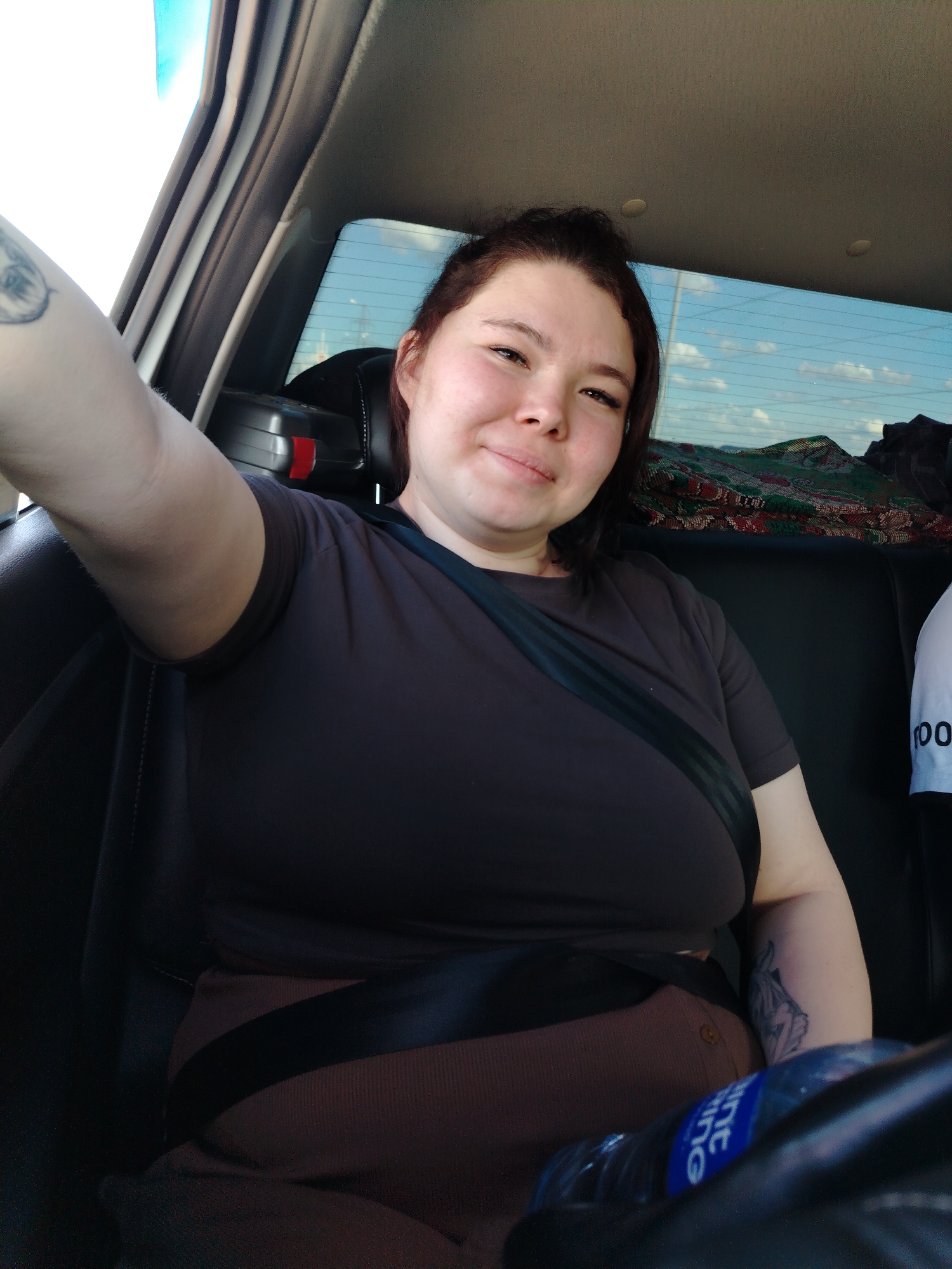 A medium shot depicts a woman in the back seat of a car. She is wearing a brown shirt and a seatbelt. She has brown hair pulled back and is looking at the camera with a slight smile. A water bottle is visible in the foreground. A blanket is on the rear deck of the vehicle. The background visible through the rear window shows a blue sky and land in the distance. It is daytime. There are tattoos on both arms of the person. This is not in Noviy, Russia.