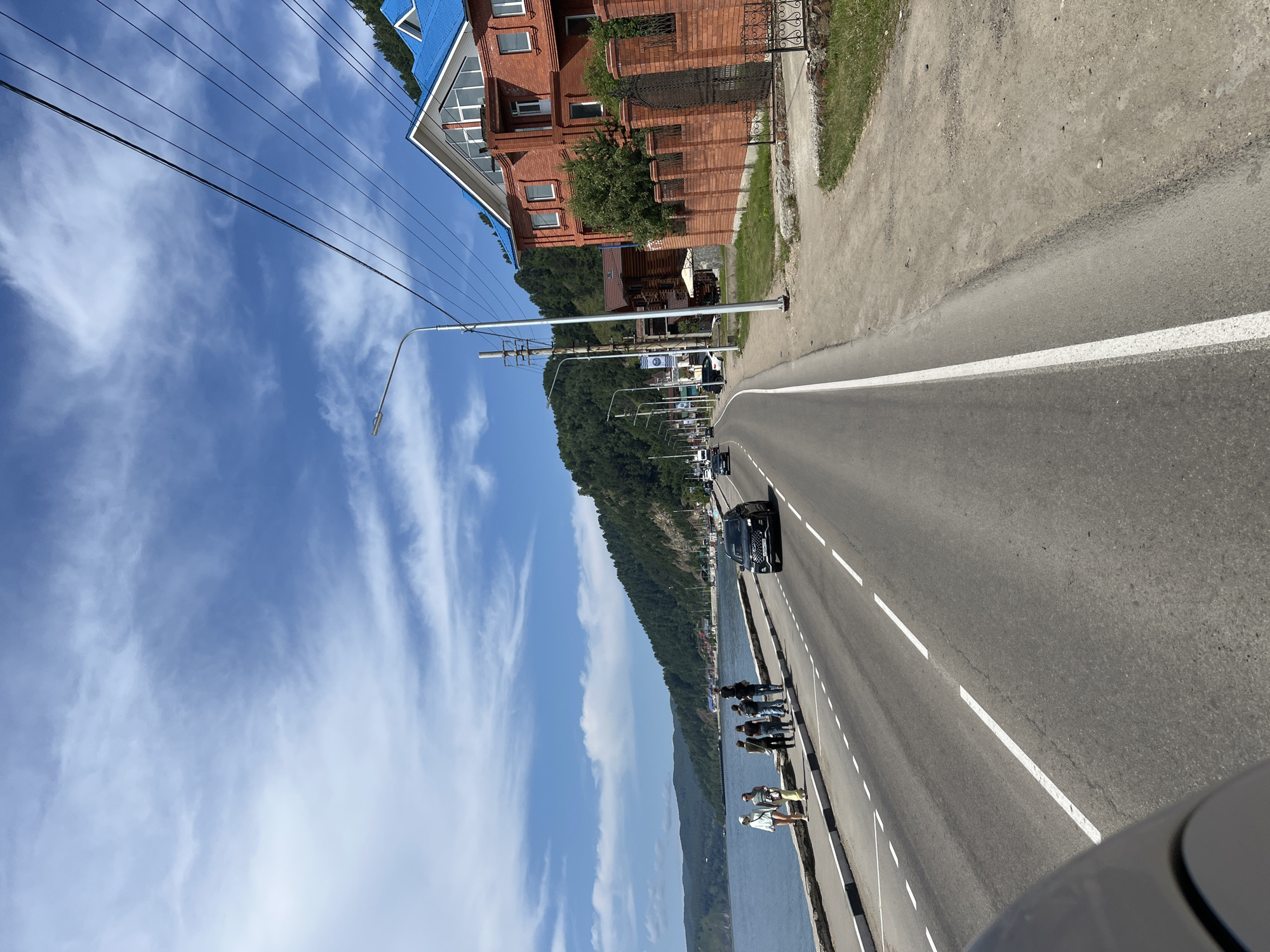 The image captures a street scene in Listvyanka, Russia, along the shore of Lake Baikal. A paved road with white dashed lane markings runs parallel to the lake. On the left side of the road, a paved walkway is occupied by several individuals, including two women wearing casual summer clothing and a group of four people, two of whom are carrying backpacks. Beyond the walkway is the calm body of water, with a tree-covered mountain range forming the backdrop.
On the right side of the road, a large brick building with a bright blue roof stands prominently. It features multiple windows and a distinctive architectural style. A low brick fence with ornate iron gates separates the property from the road. Several utility poles with numerous wires stretch across the sky above the scene. Other buildings, including a log cabin-style structure, are visible further down the road. A dark-colored SUV is driving on the road, and other vehicles are present in the distance, suggesting moderate traffic. The sky is blue with scattered white clouds.