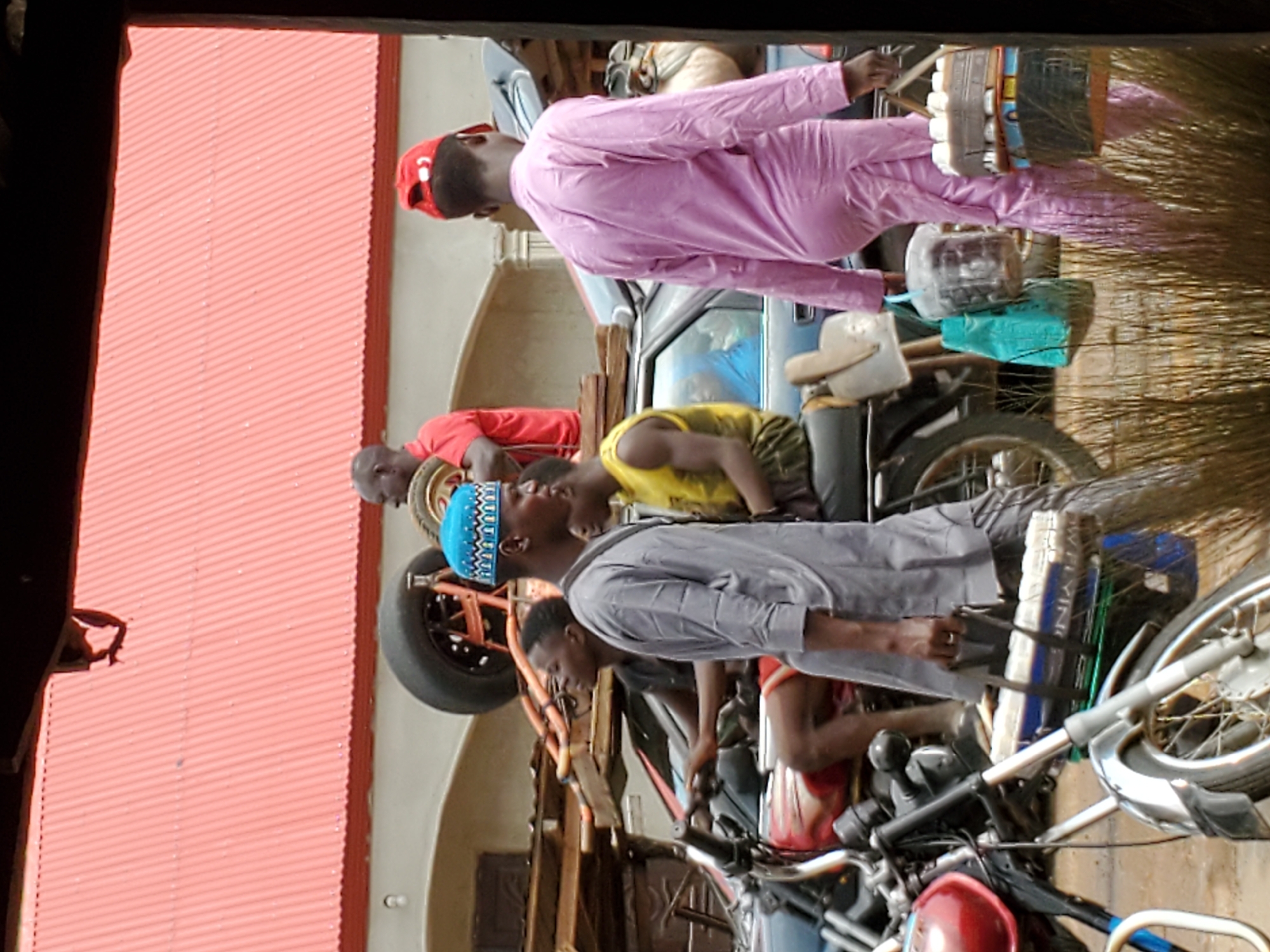 Captured in Mararaba, Nigeria, the image displays a group of males in casual attire, congregating around motorcycles. One individual is clad in pink, another in red, a third in yellow, and a fourth in grey. The scene is densely packed with motorcycles and auto parts, suggesting a mechanic shop or motorcycle repair area. A red corrugated metal roof is visible in the background. One person appears to be transporting items on a motorcycle.