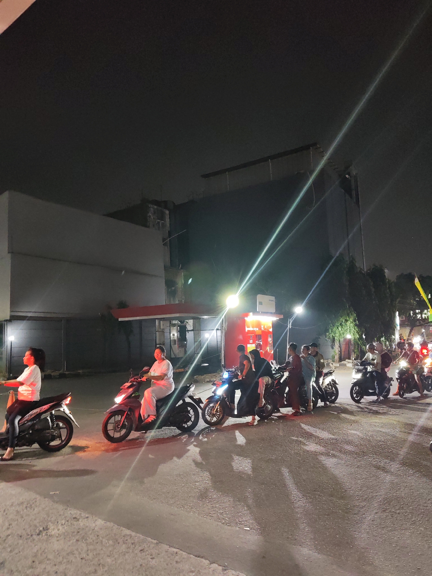 A group of people are on motorcycles at night on a street in South Jakarta, Indonesia. The lighting suggests it is evening or night, with streetlights and motorcycle headlights illuminating the scene. The people appear to be waiting or stopped, with some looking forward and others engaged in conversation. The environment consists of a paved road, buildings in the background, and some trees. The overall atmosphere is casual, with individuals dressed in everyday clothing.