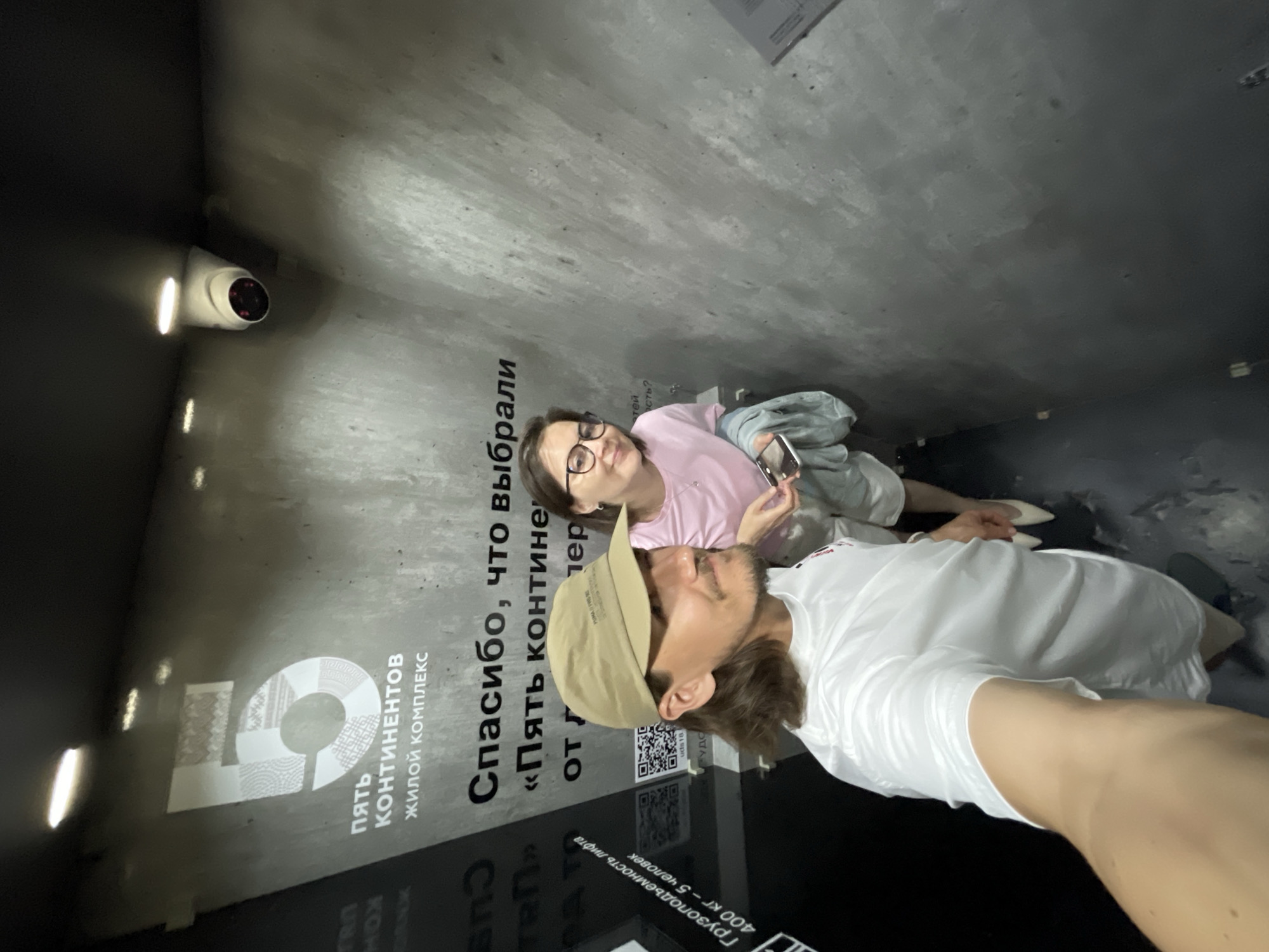 The image shows two people standing in what appears to be an elevator or small room in Izhevsk, Russia. A woman wearing glasses and a pink shirt is standing next to a man wearing a tan baseball cap and a white shirt. They are both looking towards the camera. The walls of the room are gray and have the words "Пять Континентов" (Five Continents) in Russian. There is also a security camera mounted on the wall.