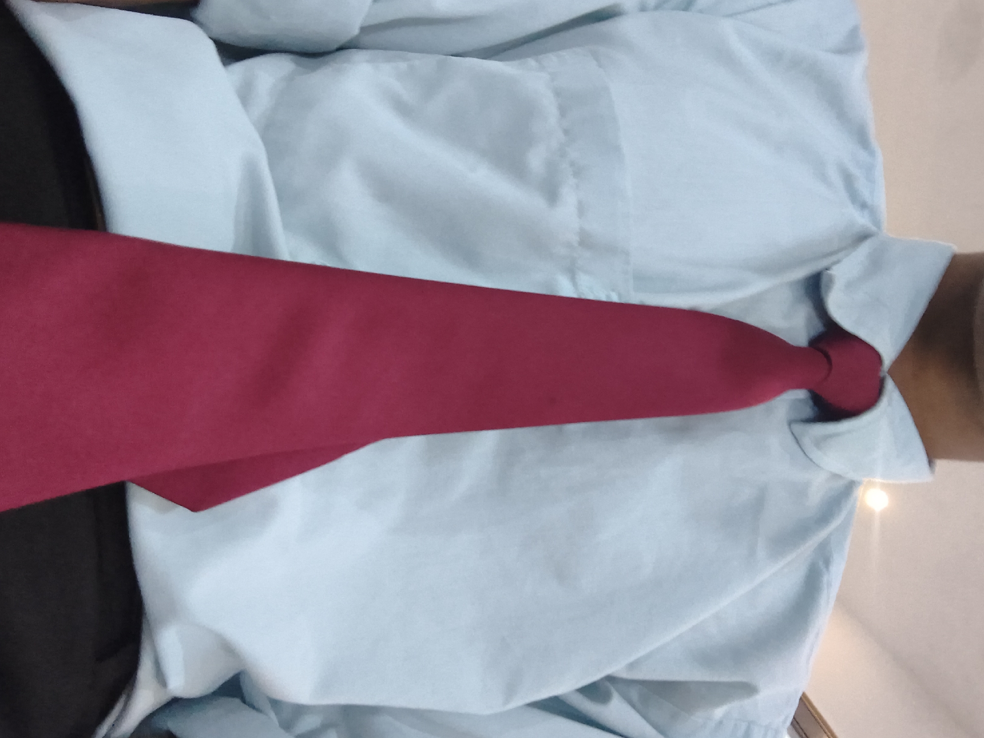 The image shows a close-up view of a person wearing a light blue collared shirt and a dark red tie. The tie is knotted at the neck and drapes down the front of the shirt. The shirt appears to be made of a light, possibly cotton, fabric. The collar of the shirt is visible, as is a portion of the person's chest and shoulders. The background is blurred but suggests an indoor setting, possibly an office or a formal event. There is no visible information about the specific location in Khaniara, India.