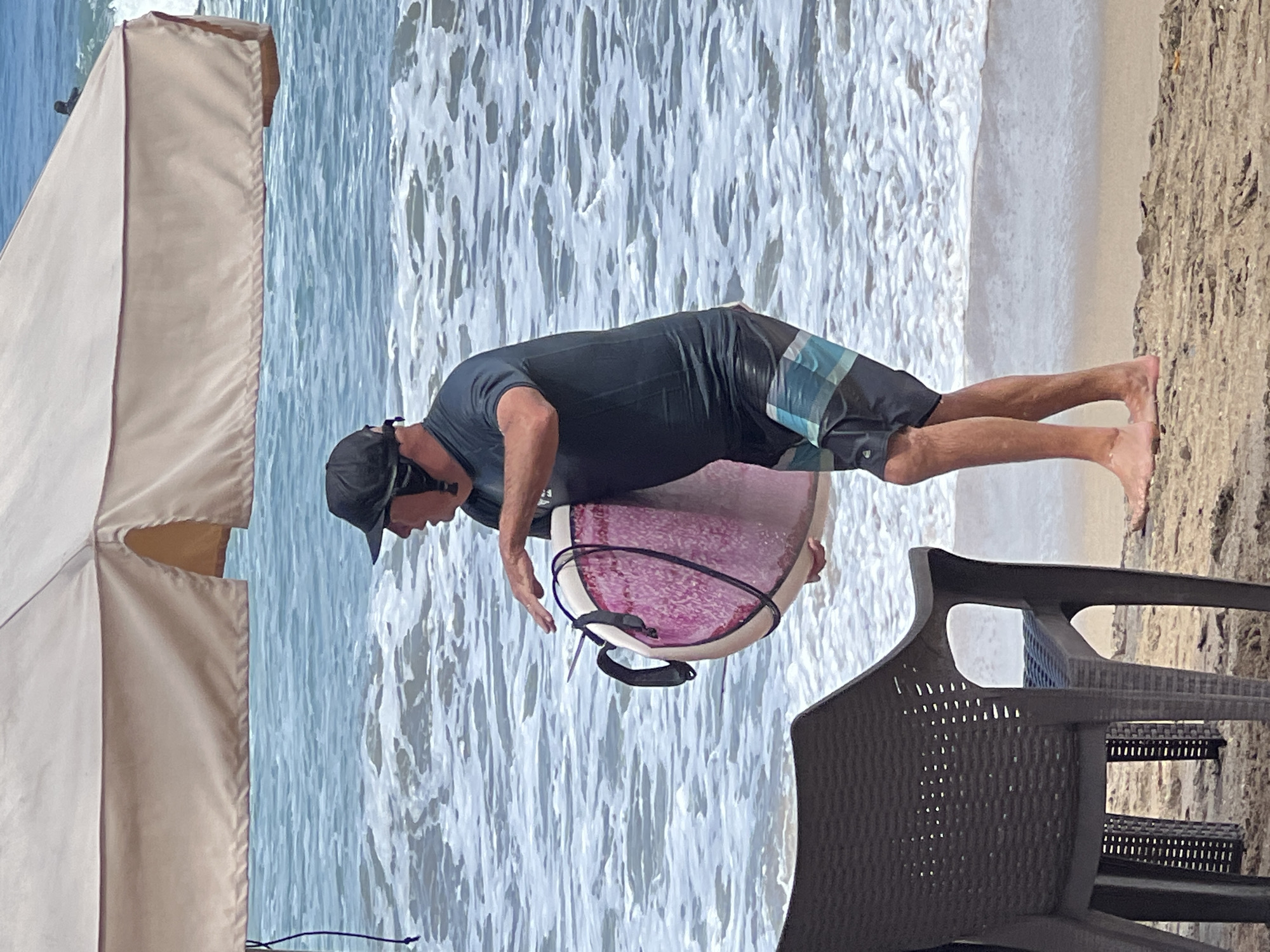 A man wearing a black cap and dark swim shorts stands on a sandy beach in Kuta, Indonesia, holding a pink and white boogie board. He is partially obscured by a large, tan beach umbrella. The ocean is visible behind him, with gentle waves lapping at the shore. A dark plastic chair is in the foreground to the left of the frame.