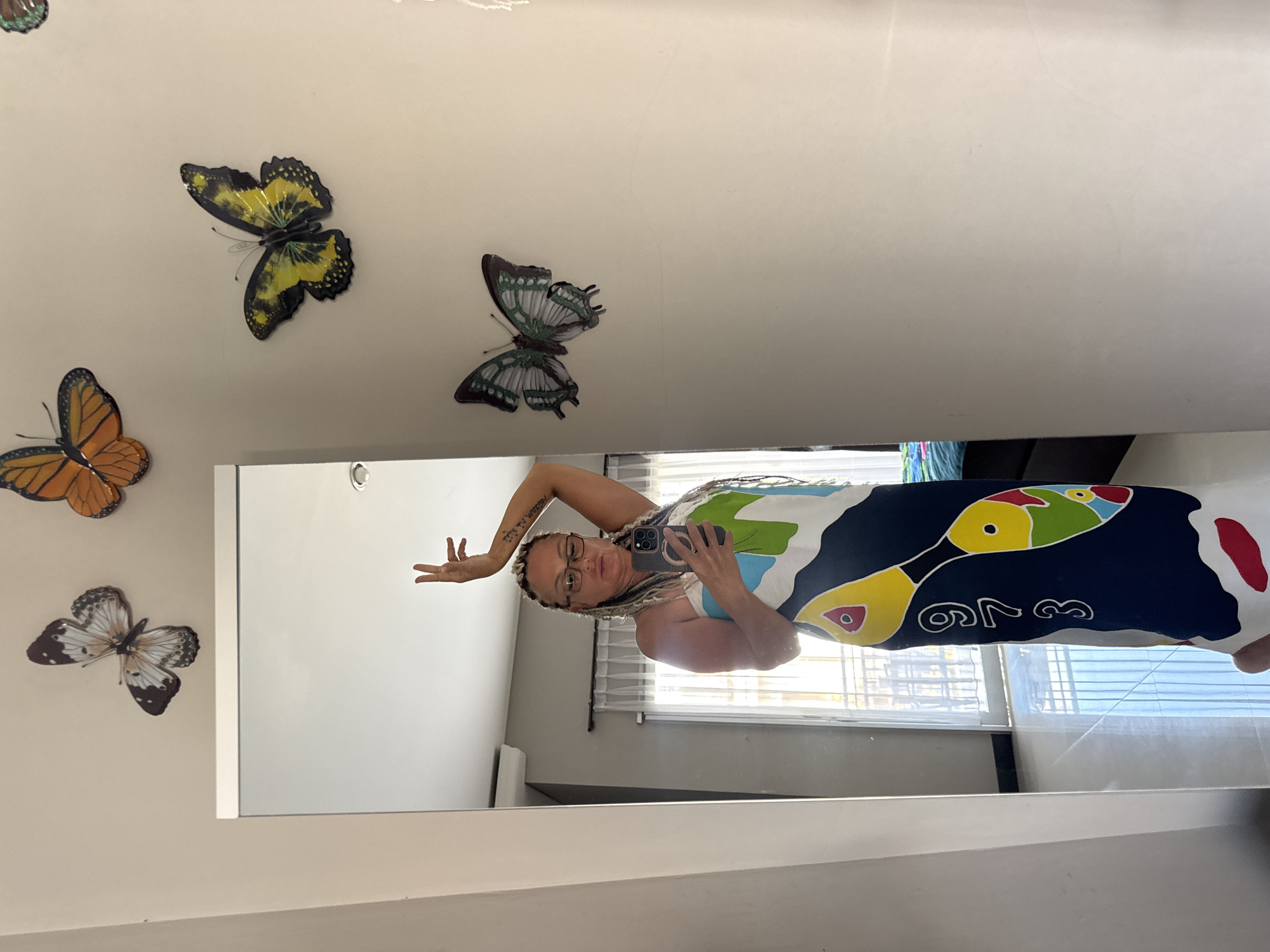 A woman with long, light-colored braids and glasses is taking a mirror selfie. She's wearing a dress with a toucan design in black, yellow, green, and red. Her left arm is raised above her head, hand open. The background is a plain wall decorated with butterfly decals in orange, yellow, and green. The reflection shows a window with light filtering through blinds. The location is likely an interior space, potentially a room in Kuta, Indonesia.
