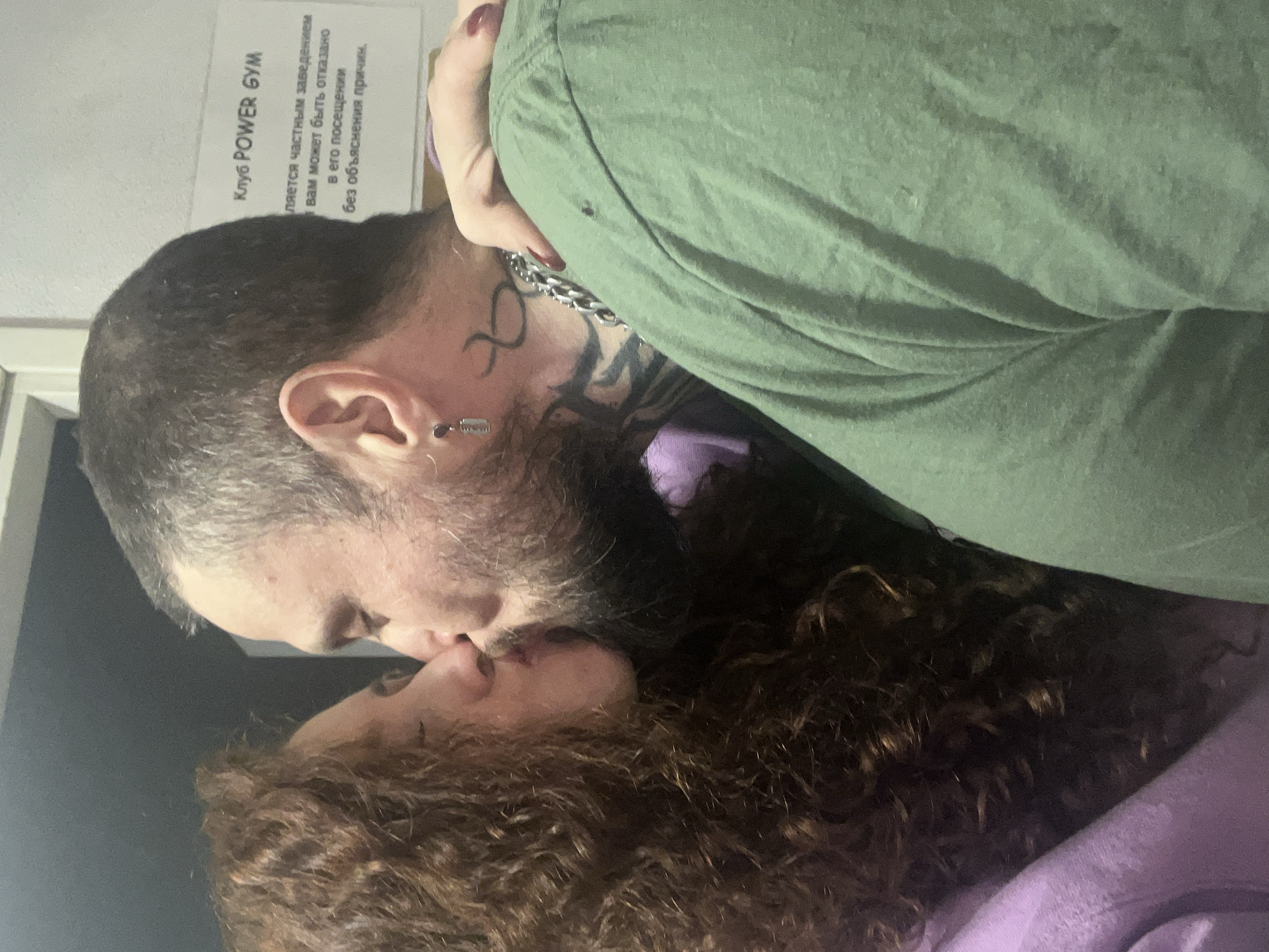 A man and a woman are kissing. The man has a beard, a tattoo on his neck, and an earring. He is wearing a green shirt and a silver chain. The woman has curly hair. The sign on the wall says "Kлуб POWER GYM" in Russian, which translates to "POWER GYM Club". The sign also states that it is a private establishment and that people may be denied access without explanation. The location appears to be in Saint Petersburg, Russia, given the Cyrillic alphabet used on the sign.