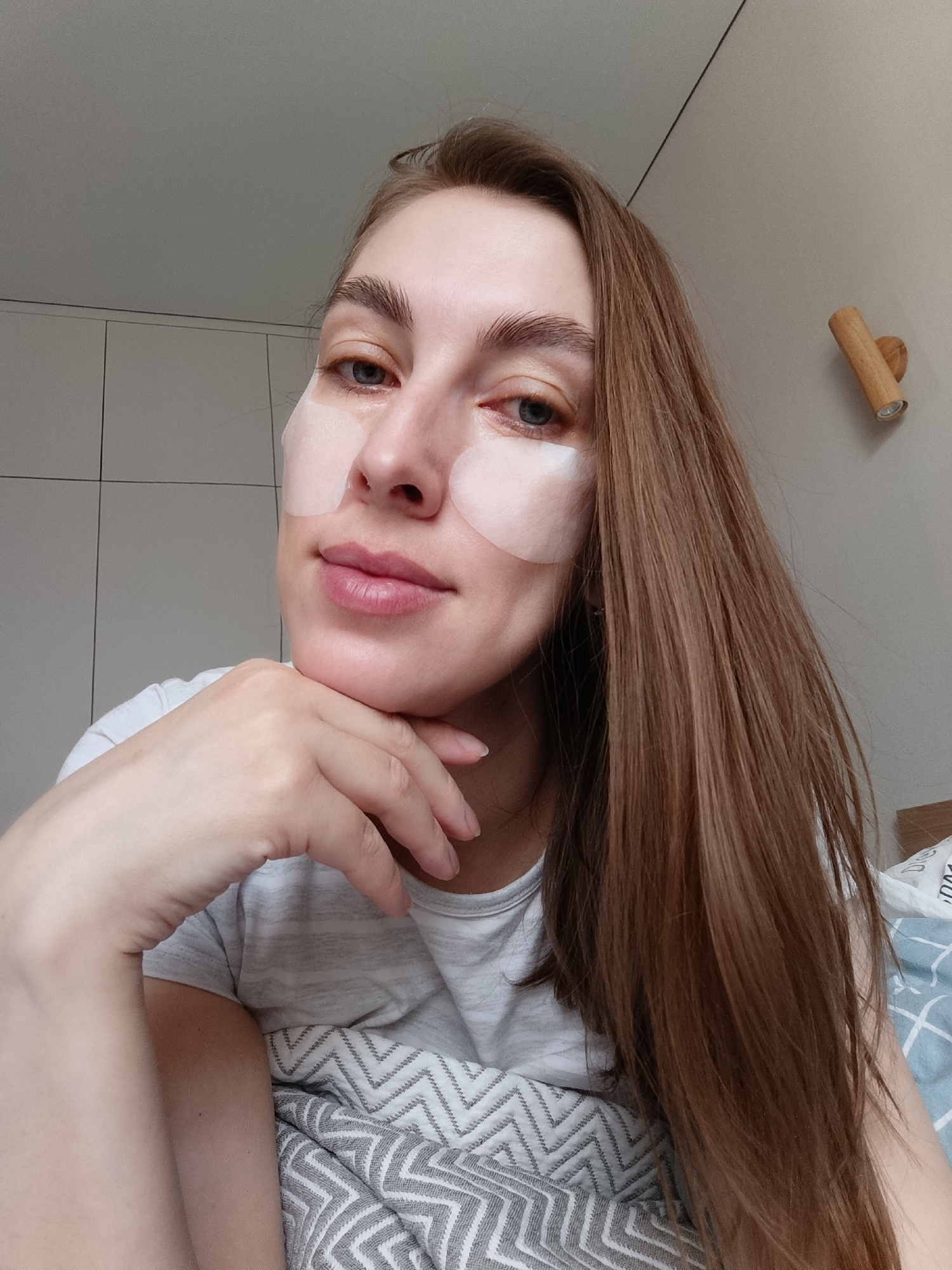 A fair-skinned woman is posing for a selfie indoors. She has straight, medium-brown hair that falls over her right shoulder. She has eye patches applied under each eye, extending onto her cheeks. The woman's face is resting on her left hand, which is in the lower left corner of the frame. She is wearing a light-gray t-shirt. The background includes a white wall with vertical and horizontal lines forming a grid pattern, possibly tiled or paneled. To the right, there is a wooden wall sconce. The lower right corner shows a bed with gray and white chevron patterned blanket. The location is Chaikovsky, Russia.