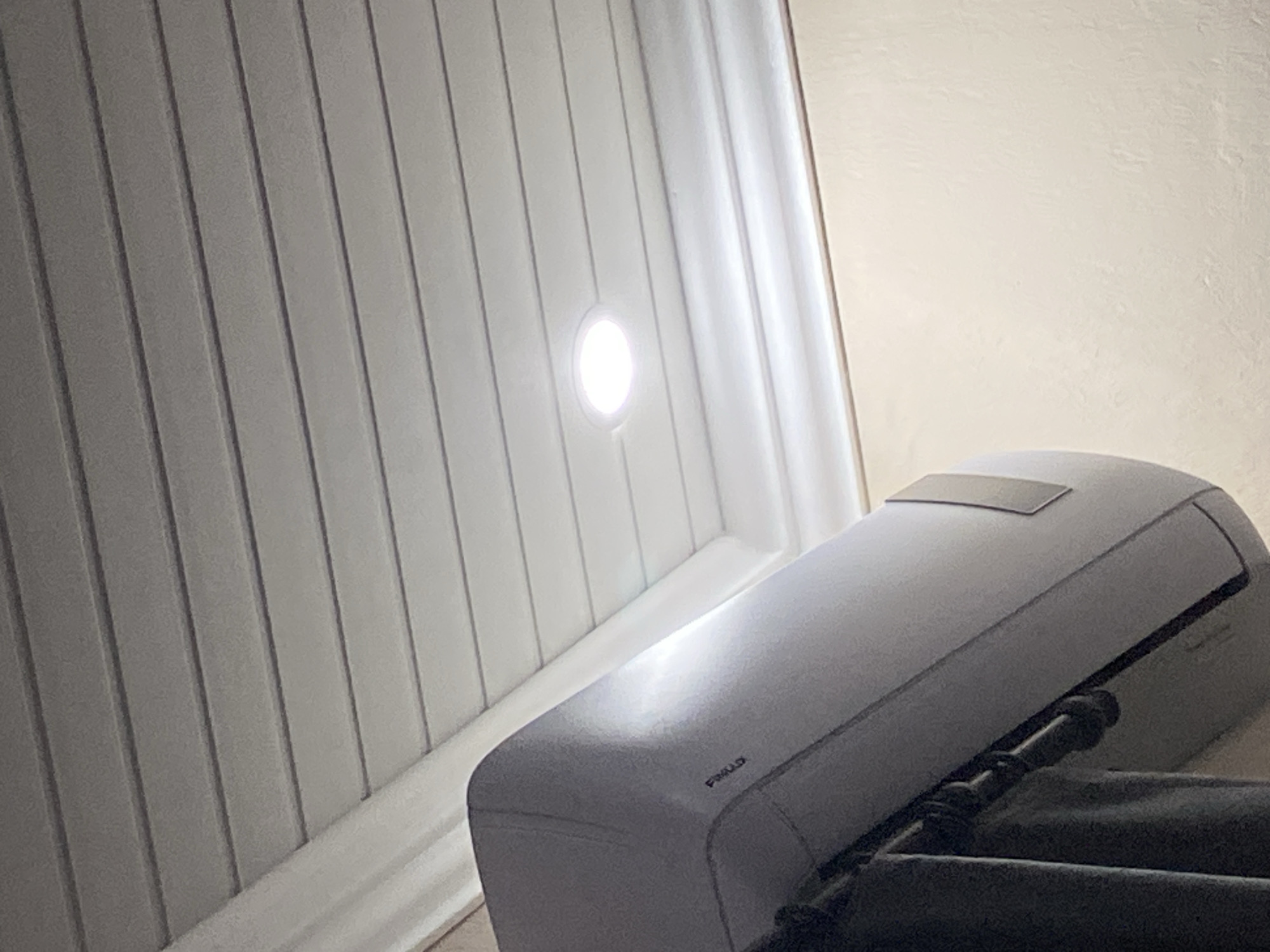 The image shows an interior scene with a white, ridged wall on the left side. A small, circular light is mounted on the wall. Adjacent to the wall is a white air conditioning unit or similar device, sitting atop a darker, possibly black, object that is partially obscured. The background on the right appears to be a textured, off-white wall. The location in the city of Konstantinovo, Bulgaria is not visually discernible from the image.