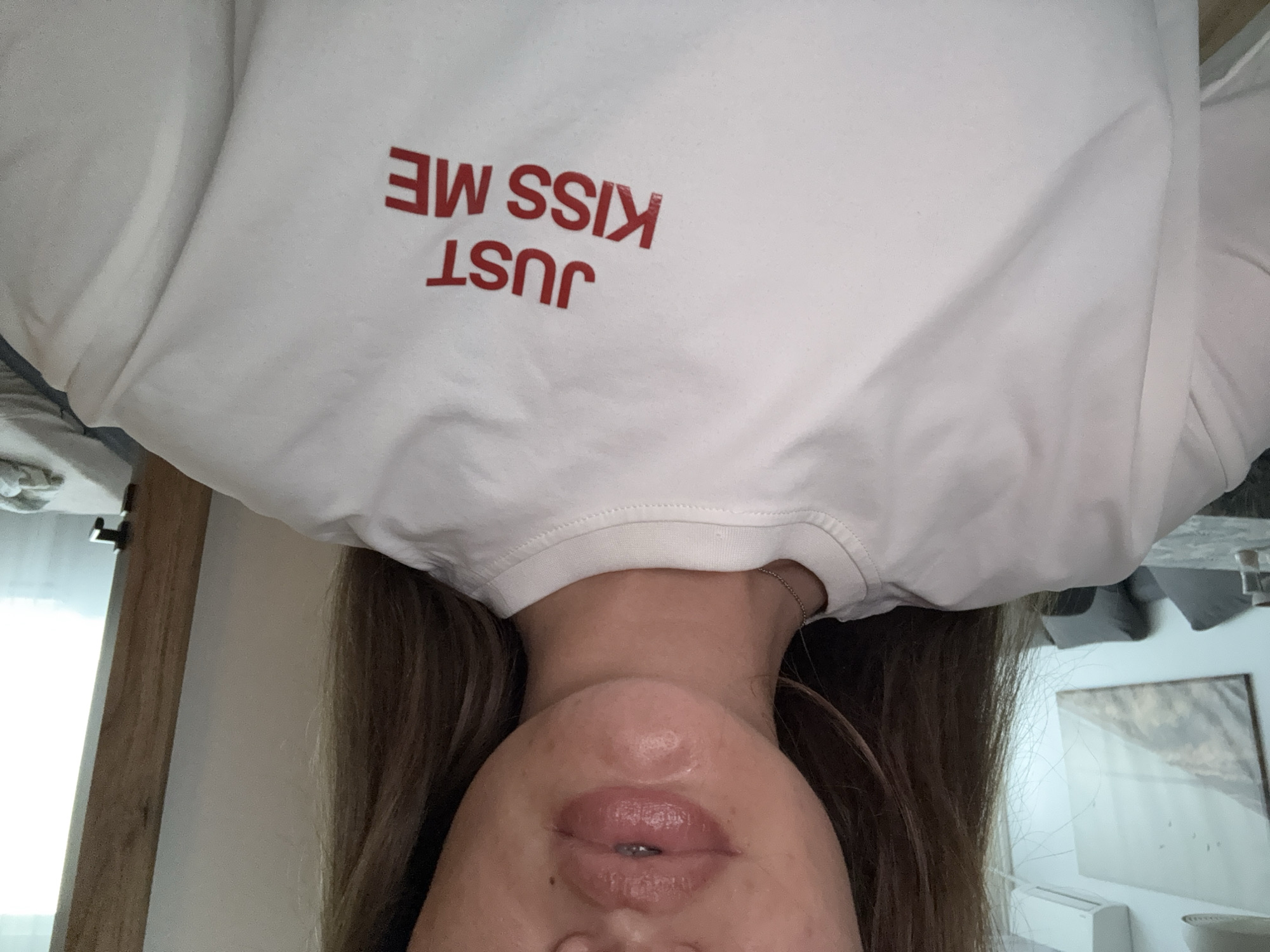 The image displays a close-up, upside-down view of a person, likely female, captured indoors. Her long, straight brown hair cascades around her head. The visible portion of her face includes her chin, full, glossy, and slightly parted lips, and the lower part of her nose, suggesting a relaxed or pouting expression. She is wearing a plain white long-sleeved top or sweatshirt. The most prominent feature of her attire is the bold red text on the front of her shirt. As it appears in the image, the text is mirrored and upside down, but when correctly oriented, it clearly reads "JUST KISS ME" in uppercase letters.
The background provides clues about the setting, suggesting a room, possibly a bedroom. To the left, a bright light source streams in from behind a vertical wooden structure, which could be a door frame or part of a wooden headboard. The main background is a simple white wall. On the right, partially visible behind the person's hair, there is a framed piece of artwork with a light-toned, possibly abstract or landscape, image hanging on the wall. Below the artwork, a white rectangular object, resembling an air conditioning unit or vent, is partially discernable. Some grey fabric, consistent with bedding or upholstery, is also visible on the far right. The lighting suggests it is daytime. The unusual orientation of the shot implies the person might be lying down while being photographed or taking a selfie.