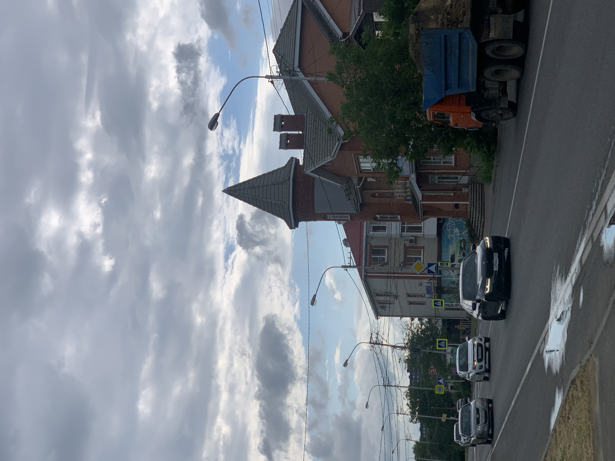 The image depicts a street scene in Stavropol, Russia, likely on a cloudy day. In the foreground, a wet asphalt road is visible, with a large puddle reflecting the sky. Several cars are driving on the road, including a black BMW in the right lane and a silver sedan in the left lane. A large dump truck loaded with dirt is moving away from the camera on the right side of the road.
In the background, there are several buildings. The most prominent is a brick building with a distinctive turret and a grey, conical roof. Adjacent to it is another brick building with a red roof. Further down the street, other buildings and trees are visible. Streetlights and power lines crisscross the sky. Several traffic signs are present, including speed limit signs and pedestrian crossing signs. The sky is filled with a mix of grey and white clouds, with patches of blue sky visible. There are no visible people in the image.