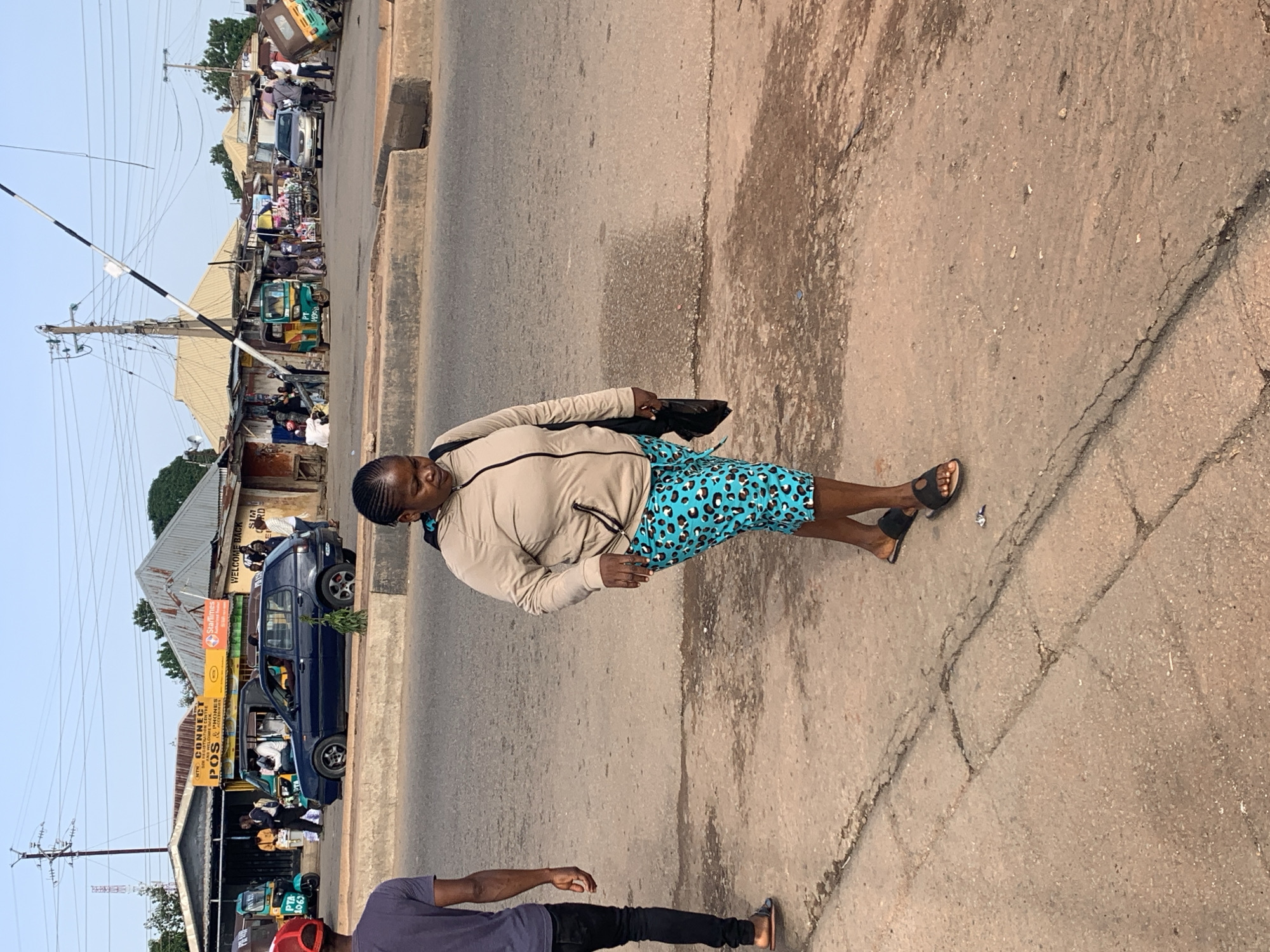The image depicts a street scene in Gura Top, Nigeria, likely in the late afternoon or early evening, judging by the soft lighting and long shadows. A woman wearing a light beige jacket and blue and black leopard-print shorts stands in the foreground, facing towards the right side of the frame. She carries a dark bag over her shoulder.
Behind her, the street is lined with various shops and stalls. Several vehicles are visible, including a dark blue car with tinted windows and a three-wheeled auto-rickshaw. People can be seen gathered around these vehicles and near the shops, suggesting a busy commercial area. Power lines and utility poles stretch across the upper part of the scene, typical of urban or semi-urban African landscapes.
One shop has a prominent yellow sign with black lettering that reads "CONNECT", followed by "POS & PHONES". Another sign, partially visible, displays "WELCOME BACK". The overall impression is one of a bustling street market or commercial strip. The ground is paved with asphalt and shows signs of wear and tear, with visible cracks. The weather appears to be clear.