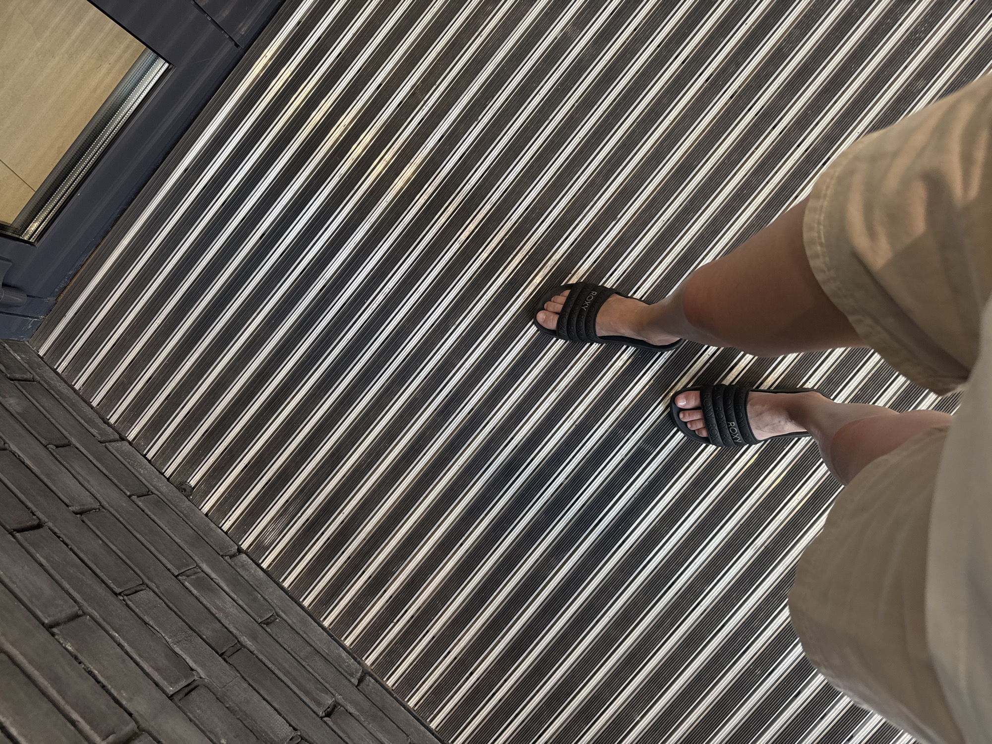 The image shows a first-person perspective of a person standing on a striped mat. The mat has alternating black and silver stripes running diagonally from the bottom left to the top right of the frame. To the left of the mat is a brick wall constructed with dark grey bricks laid horizontally. In the upper right corner, a section of a doorway with a dark grey frame and glass is visible, with a light-colored floor beyond it. The person is wearing light-colored shorts and black slide sandals. One sandal is clearly visible on the right foot, and the left sandal is partially visible. The person's legs and feet are positioned in the lower half of the image, with the feet pointing towards the top right. The scene appears to be an indoor or covered outdoor entryway, possibly a porch or a hallway, given the presence of a doormat and a door frame. No specific geographical markers indicate the location in Krasnoyarsk, Russia.