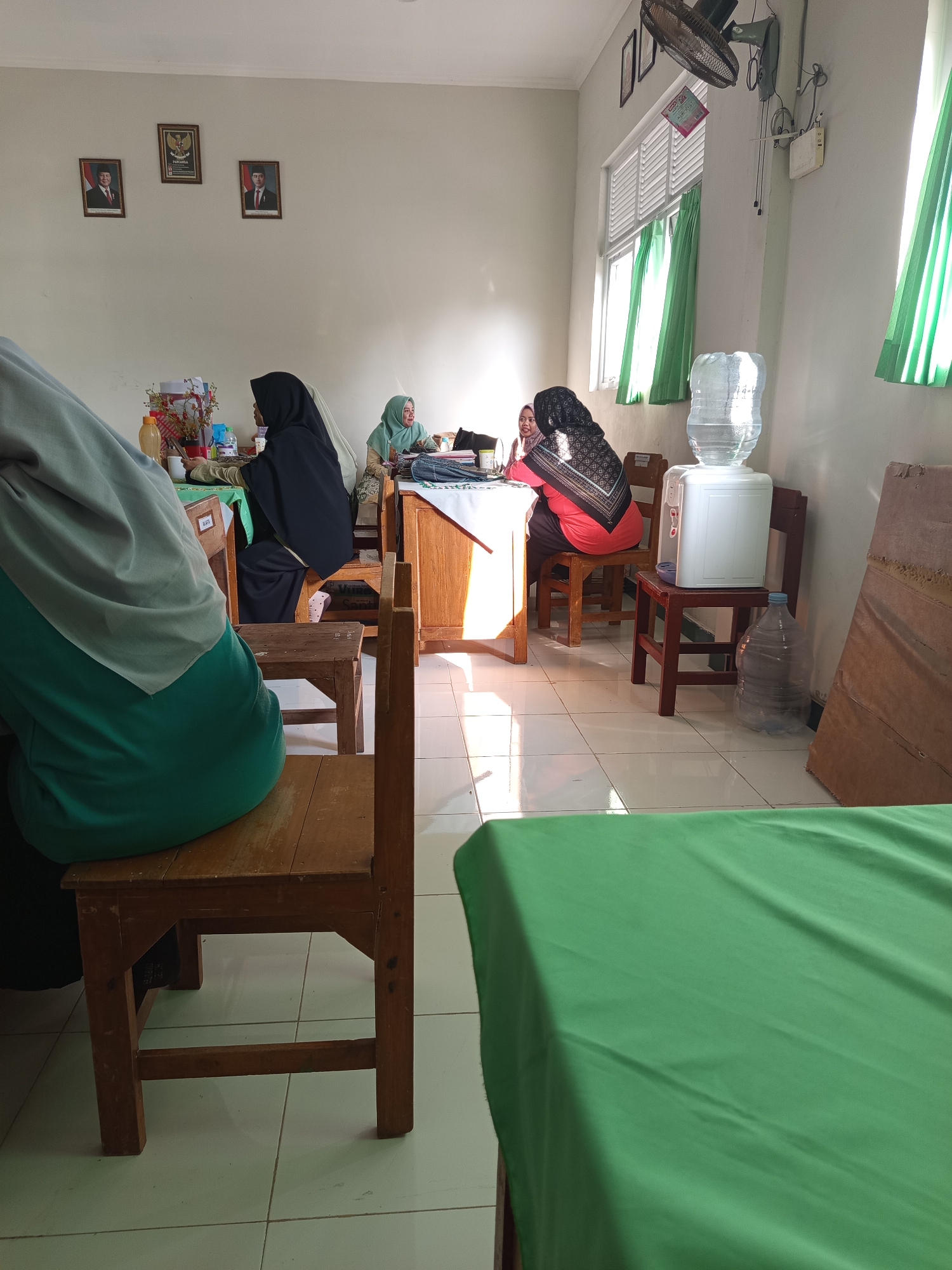 The image depicts an indoor scene, likely a classroom or office, with natural light streaming in from windows on the right. In the foreground, a person wearing a grey and green hijab is seated on a wooden chair, their back to the camera. In the middle ground, three women are seated at a table. The woman on the left wears a black hijab, the woman in the center wears a green hijab and appears to be speaking, and the woman on the right wears a red top and patterned hijab. A water cooler with a large bottle of water is visible on a stand to the right of the table. The room has tiled floors and plain walls, adorned with framed pictures of men. Green curtains hang over the windows. The overall impression is of a quiet, perhaps administrative, setting. The location is Lumutan, Indonesia.