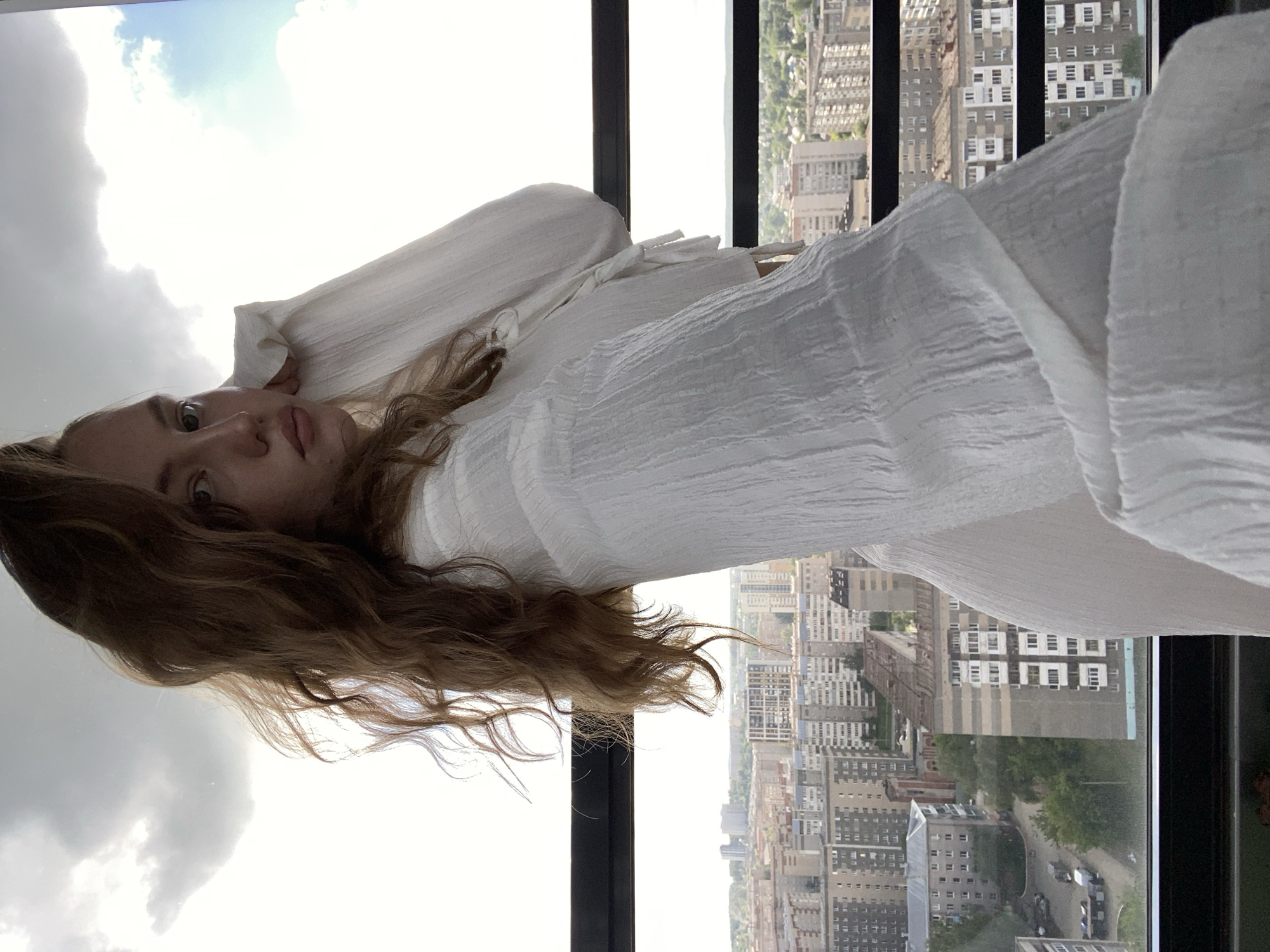 A young woman with long brown hair is wearing a white, textured dress and is positioned in front of a tall window with black frames. The window provides a view of a cityscape. Based on the architecture visible in the background, the location is identified as Izhevsk, Russia. The woman's head is tilted towards the left side of the frame, and she is looking towards the camera. Outside the window, a sky with light clouds is partially visible.