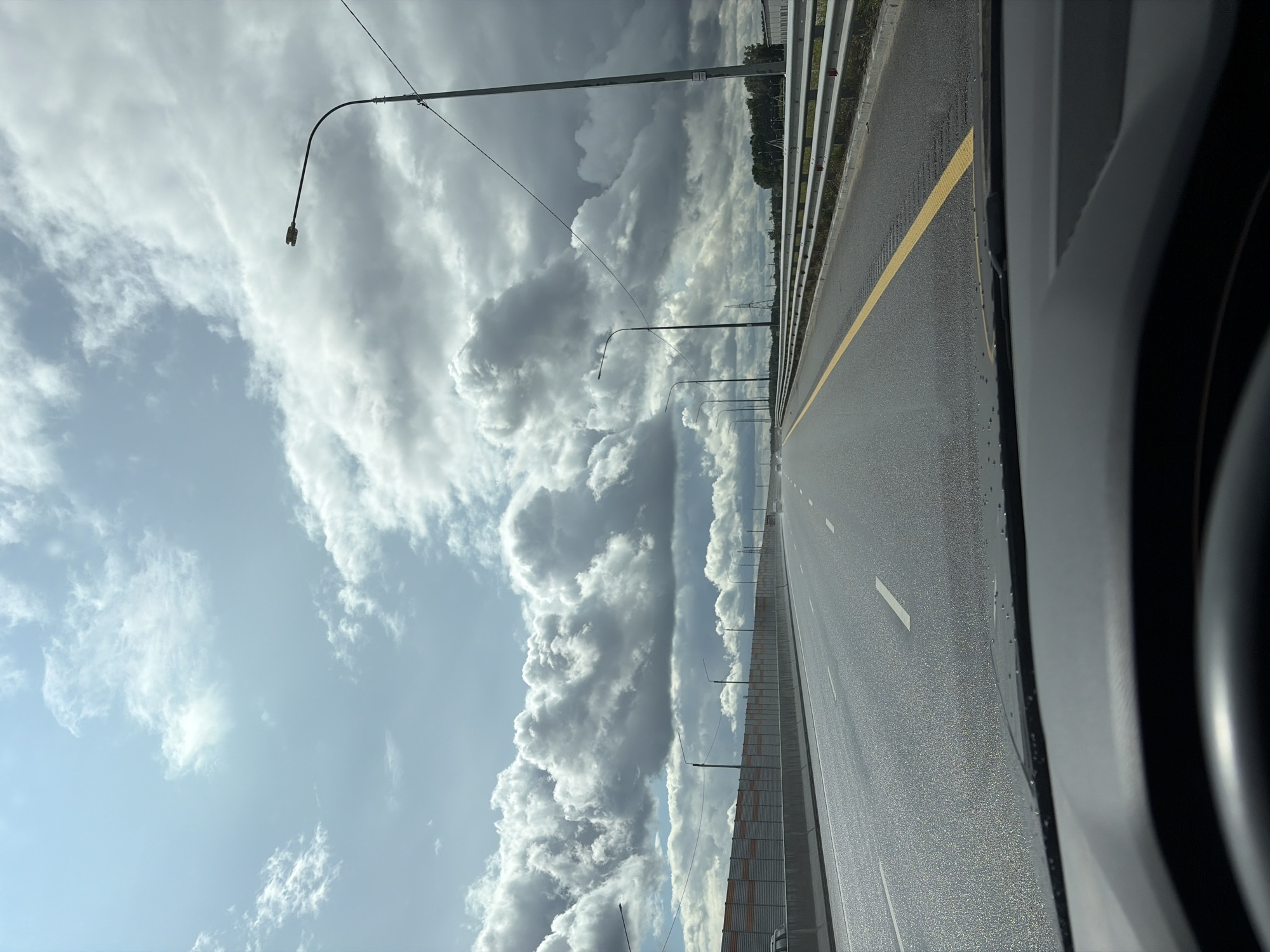The image is a first-person perspective from inside a vehicle, looking out at a multi-lane highway. The road is paved and wet, reflecting the bright sky. White lane markings are visible, along with a solid yellow line dividing the lanes. Guardrails line the right side of the highway. On the left, a sound barrier wall with orange accents separates the highway from what appears to be an undeveloped area or construction site. The sky is dramatic, filled with heavy, dark clouds mixed with patches of bright blue sky. Several streetlights are visible along the side of the road. In the distance, trees and some industrial buildings can be seen. The overall impression is that of a highway on a partly cloudy day, possibly in an industrial or suburban area on the outskirts of a city. The specific location within Balashikha, Russia, cannot be determined from the visual information alone.