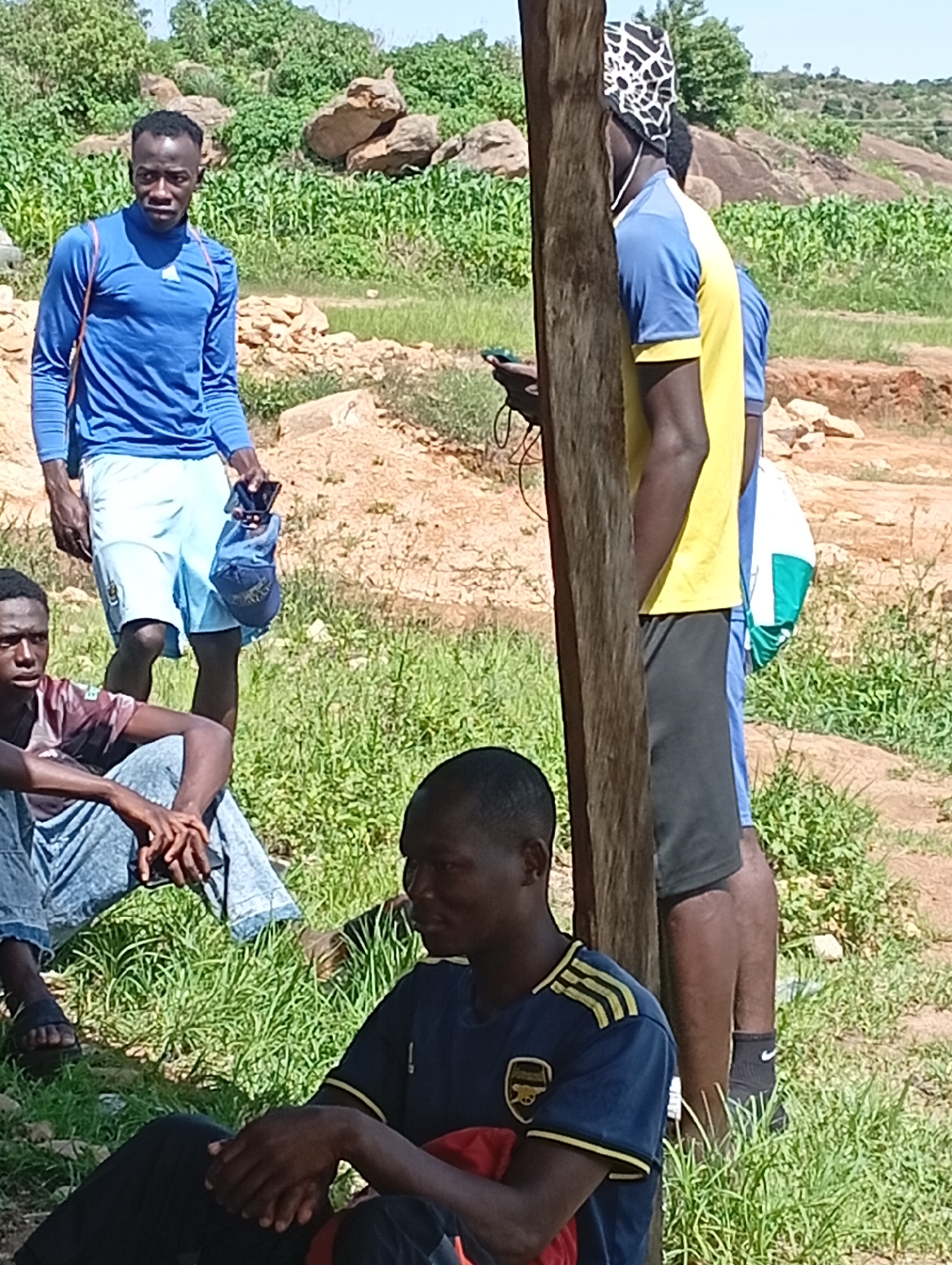 The image depicts a group of young men in an outdoor, rural environment, likely on the outskirts of Jos, Nigeria. In the foreground, two men are seated in the grass, one wearing a dark blue t-shirt with yellow accents and an Arsenal logo, the other partially visible with a burgundy and maroon t-shirt. Behind them, a taller man stands wearing a blue long-sleeved shirt and light blue shorts, holding a blue cap and a mobile phone. To his right, another man stands wearing a yellow t-shirt with blue sleeves and a black headscarf. The background features a field of green crops, rocky outcrops, and sparse vegetation under a bright, clear sky. The terrain appears uneven and somewhat arid, characteristic of the plateau region. The overall impression is a casual gathering in a semi-agricultural setting.