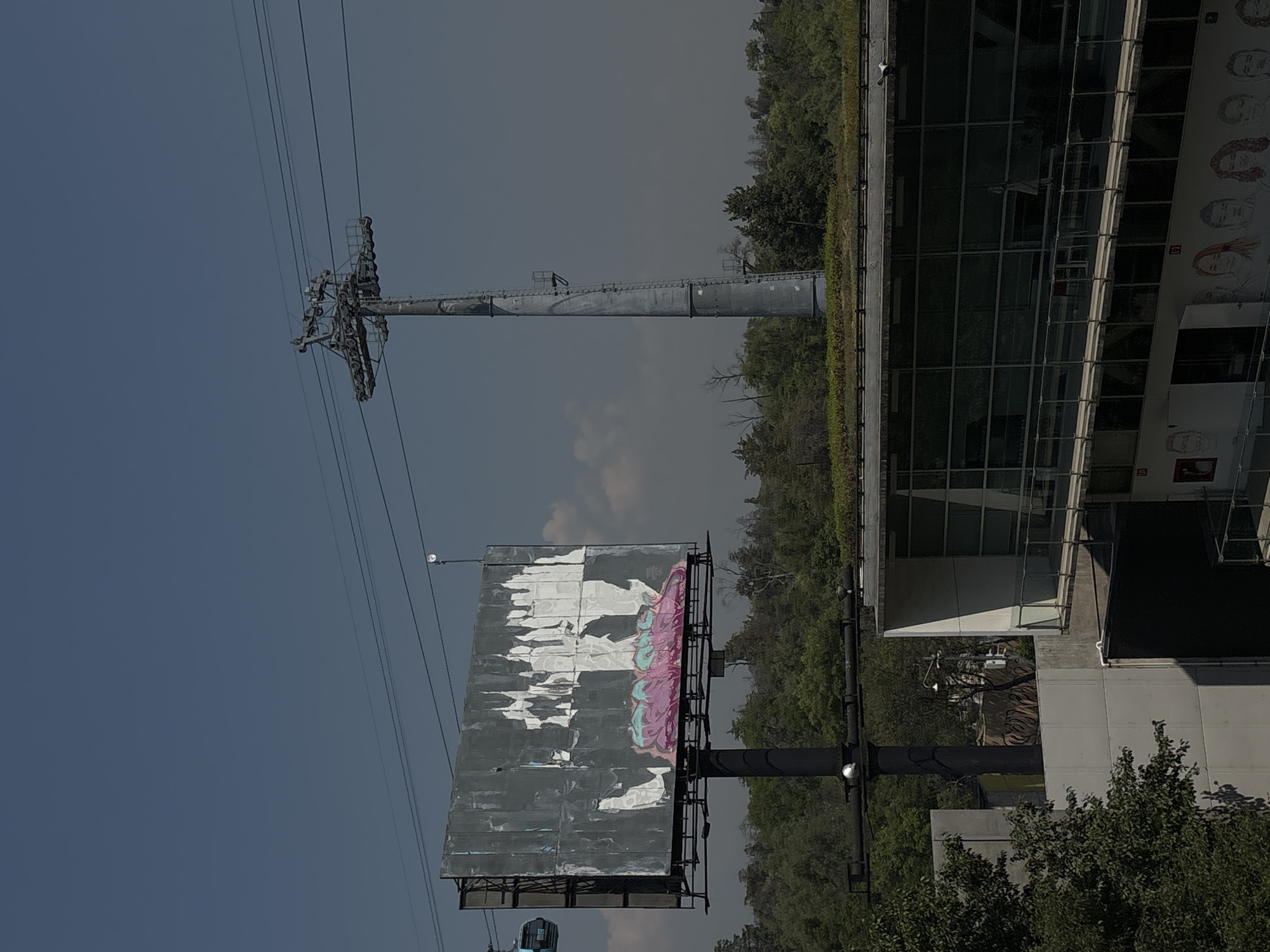 The image depicts an outdoor scene in Mexico City, Mexico. On the left, a weathered billboard displays remnants of old advertisements, with peeling paint and faded colors. Adjacent to the billboard is a cable car tower, supporting overhead cables. To the right, a modern building with large glass windows and a green rooftop is visible. The scene is set against a backdrop of trees and a somewhat hazy sky.