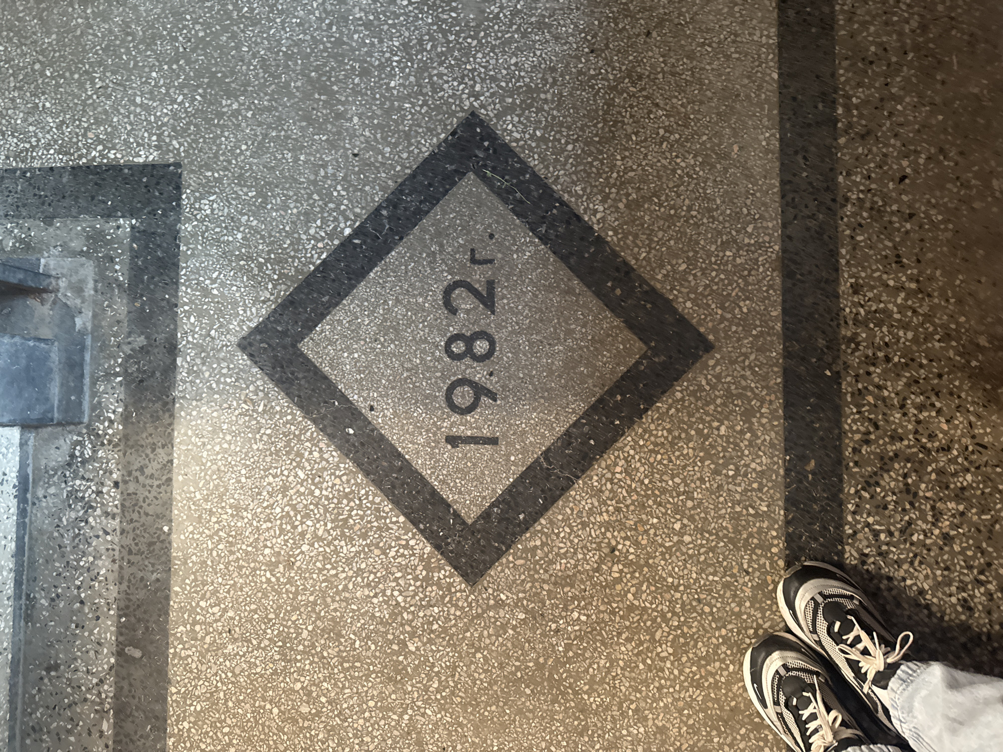 The image displays a top-down view of a textured floor, likely terrazzo or an aggregate concrete, featuring a decorative inlay. Centrally positioned is a dark grey/black diamond shape set into the lighter speckled flooring. Inside this diamond, the year "1982 r." is prominently marked in black lettering, oriented vertically. The "r." signifies "year" (година) in Bulgarian, aligning with the provided context of Montana, Bulgaria.
The diamond design is bordered by a wider, dark grey/black rectangular strip that runs along the left and right sides of the visible floor, suggesting a pathway or a larger floor pattern. In the bottom right corner, a pair of feet wearing black and white athletic sneakers are partially visible, indicating a person is standing on the floor and capturing the image. The lighting appears to be artificial and internal, without any cues regarding the time of day or outdoor weather. The overall scene suggests an interior space within a public or institutional building, where the floor design might commemorate a specific year related to the building's construction or significance.