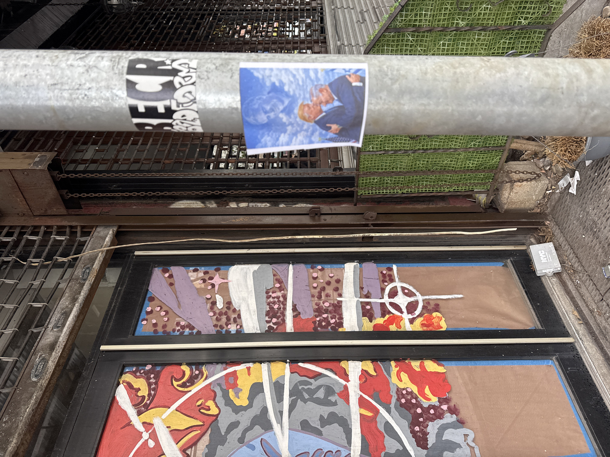 This image shows a street scene in New York City, United States of America. A metal pipe runs horizontally across the top of the frame, with two stickers adhered to it. One sticker is black and white with abstract designs and lettering. The other sticker is a color image depicting former presidents George W. Bush and Barack Obama embracing, set against a blue-toned backdrop. Below the pipe are two black-framed windows with colorful, unfinished painted designs. The designs include red, yellow, gray, white, purple, and brown elements, with geometric shapes and what appear to be partial figures or symbols. The environment appears urban and industrial, with metal grating visible both above and to the side of the windows. There are also patches of greenery and some dried leaves or debris. A small gray box is mounted to the right of the windows.