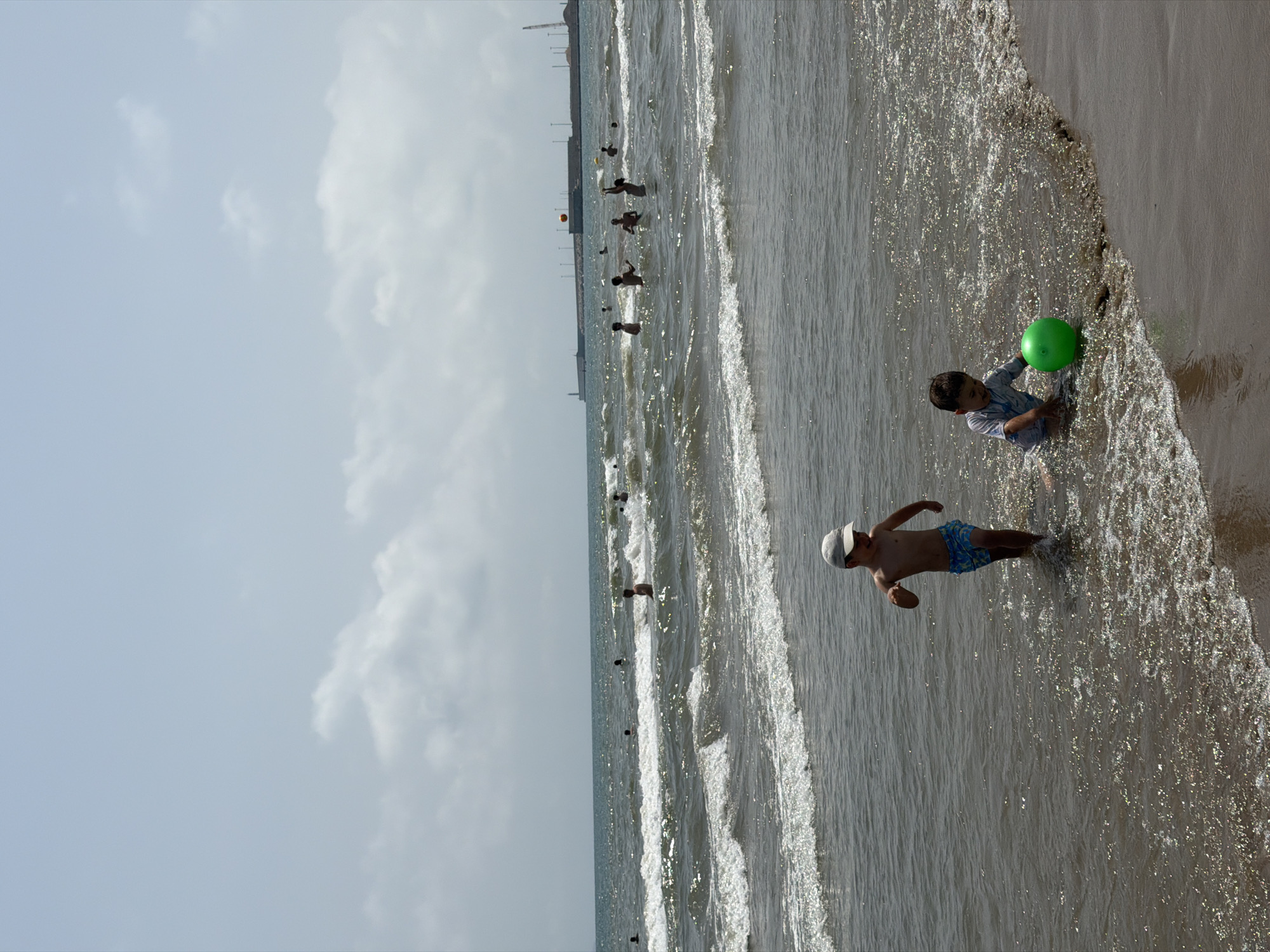 The image depicts a sunny day at a beach. In the foreground, two young boys are playing in the shallow water. The boy on the left, wearing a white cap and blue patterned swim trunks, is running towards the right. The boy on the right is sitting in the water and playing with a green ball. The sea is calm with gentle waves, and in the background, several other people are visible swimming and enjoying the water. Beyond the sea, a distant shoreline with some structures, possibly a port or industrial area, and a crane can be seen under a cloudy sky. The lighting suggests it is daytime.