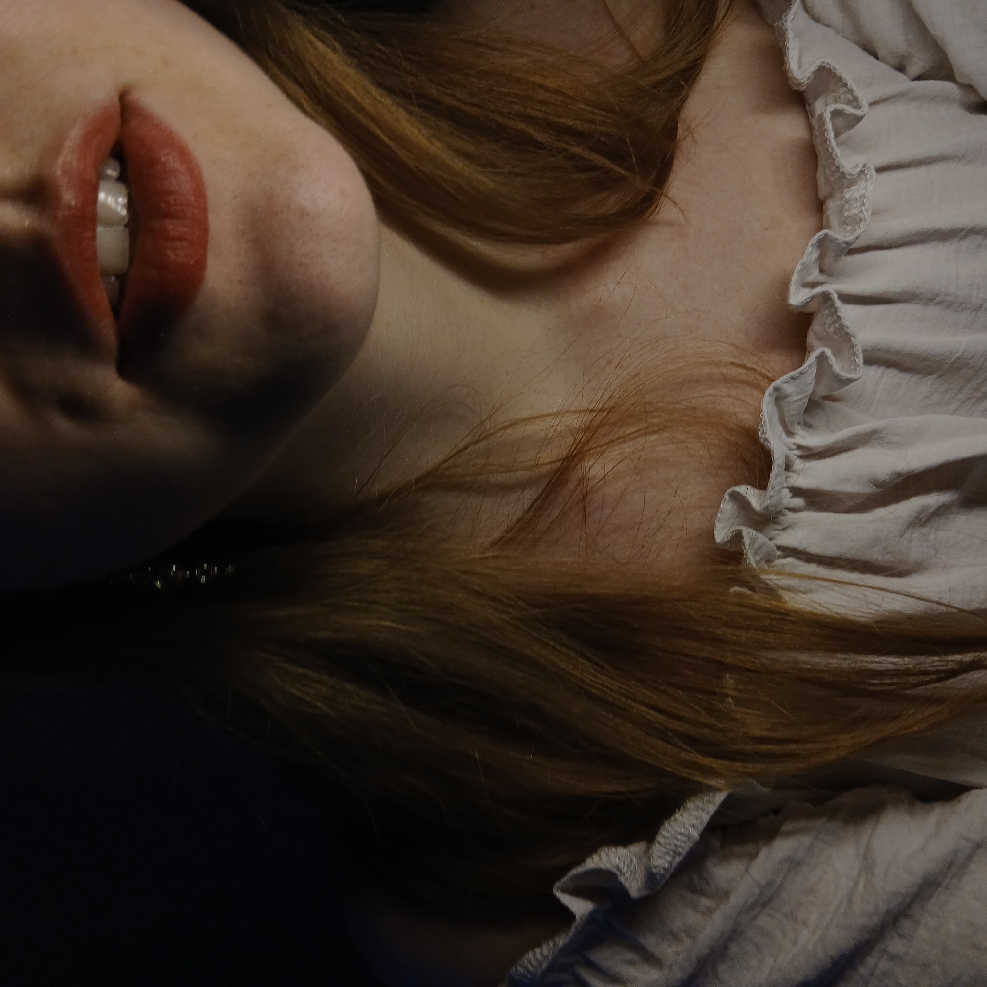 The image presents a close-up, intimate view of a person, likely a woman, angled from her chin and mouth down to her upper chest. Her lips are painted with a reddish-orange lipstick, slightly parted to reveal a glimpse of her white teeth, suggesting a subtle smile or gentle expression. Fair skin with visible freckles is present around her mouth and chin. Long, reddish-brown hair flows over her shoulder and chest, appearing thick and slightly wavy.
She is wearing a light-colored garment, possibly white or off-white, characterized by ruffled detailing along what appears to be a neckline or trim, resting near her collarbone. A delicate shimmer of small, light-colored, possibly pearl-like beads is faintly visible under her chin, suggesting a necklace or other jewelry. The background is dark and out of focus, putting the subject clearly in focus. The lighting is soft and somewhat dim, casting gentle shadows that define the contours of her face and neck. The overall impression is one of serenity and gentle beauty. No specific activity is depicted beyond this pose. There is no visible text in the image. The setting appears to be indoors, though no specific location cues are present.