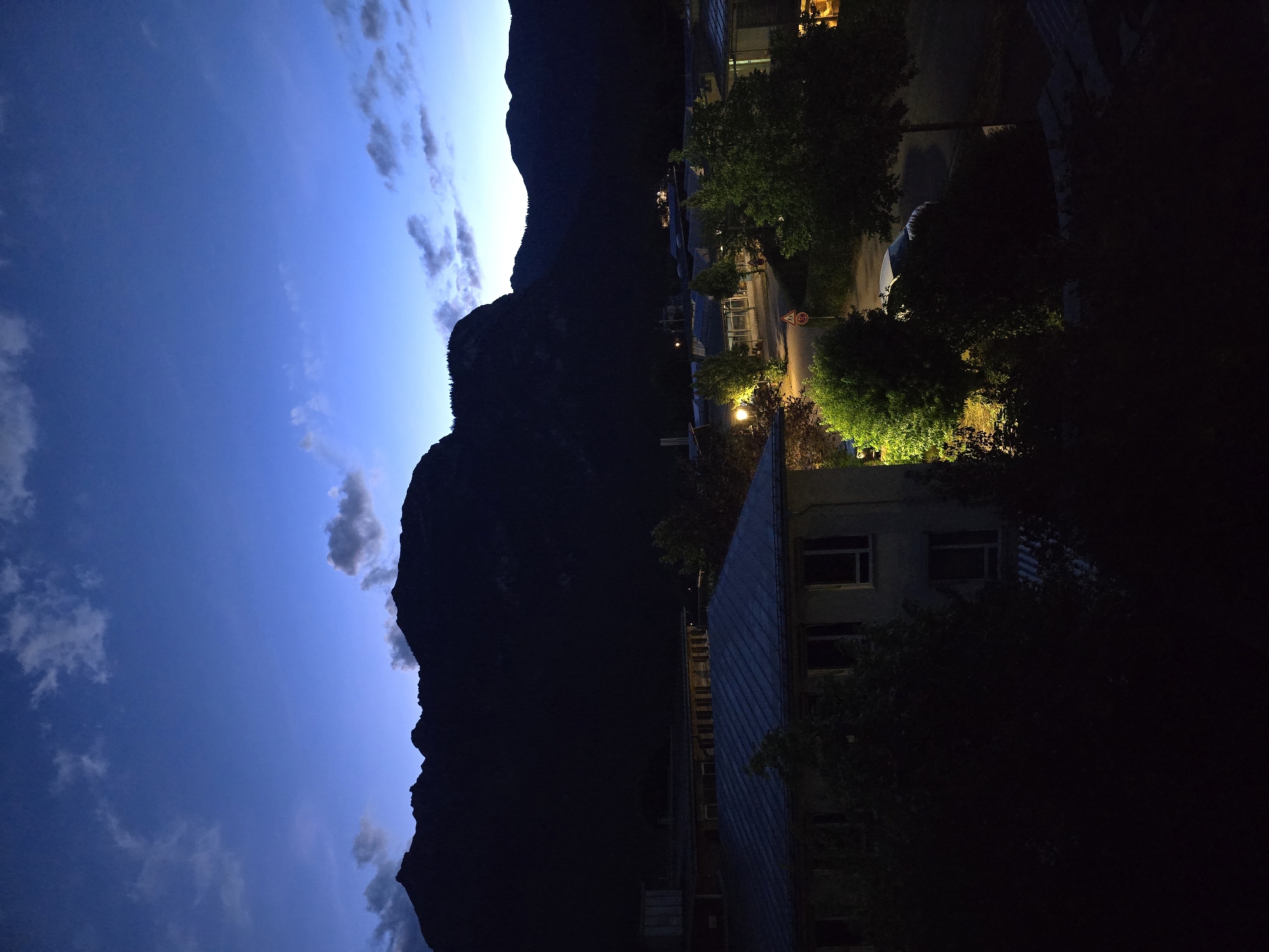 This image depicts a nighttime scene in Garmisch-Partenkirchen, Germany. The foreground features a building with a dark, sloped roof and windows. Several trees with dense foliage are situated around the building and in the mid-ground, some illuminated by artificial lights. A road or pathway is visible, with a traffic sign and a parked car. In the background, a vast mountain range forms a dark silhouette against a deep blue twilight sky with scattered clouds. The overall impression is a peaceful, mountainous town at dusk or dawn.