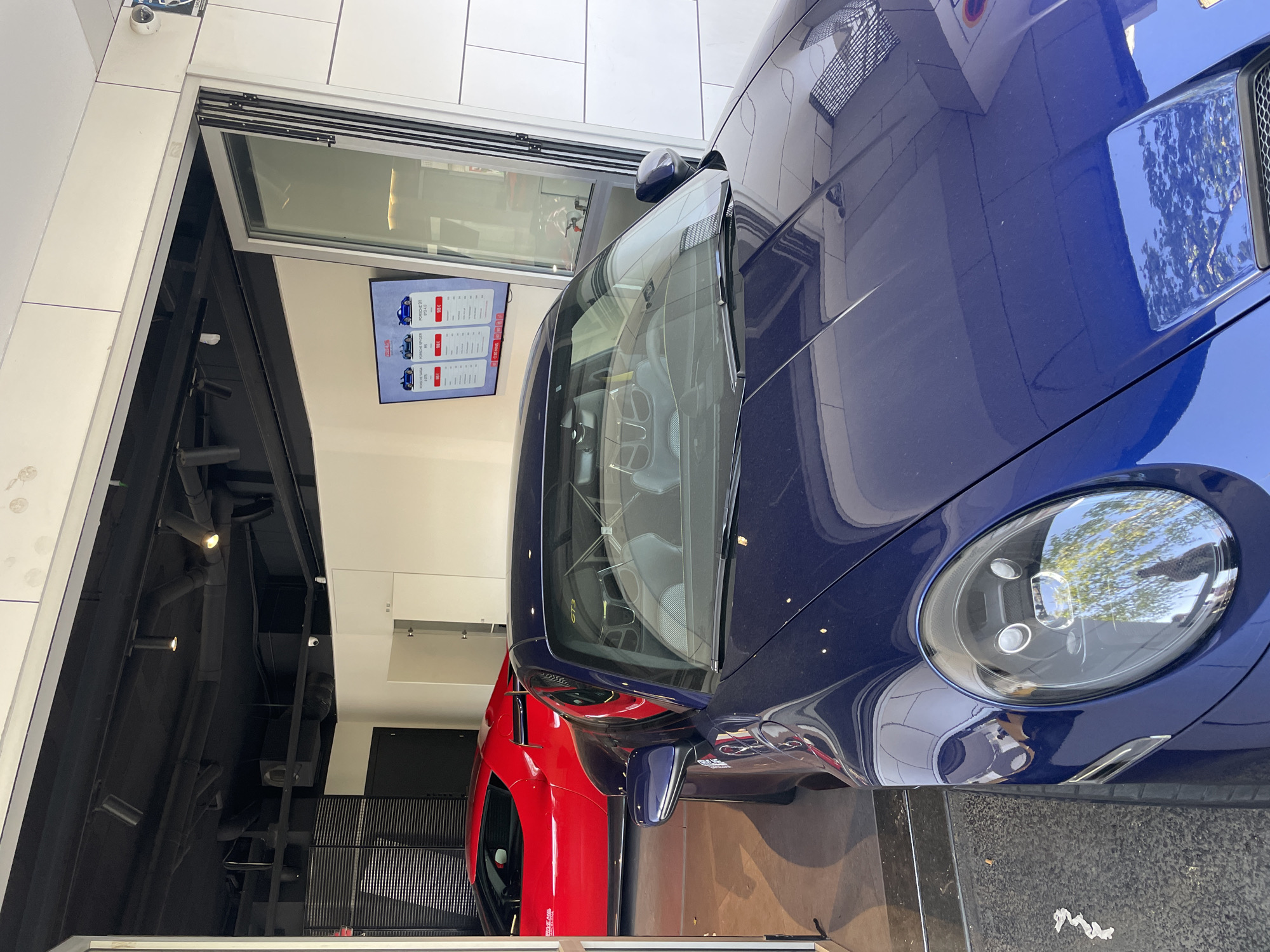A slanted, medium-shot photograph shows a blue Porsche 911 GT3 car in the foreground and a red Ferrari in the background, presumably in a dealership in Barcelona, Spain. The blue Porsche takes up a significant portion of the frame, with its headlight prominently displayed. A red Ferrari is visible behind the Porsche. There is a wall with a monitor displaying car options and an open view towards what appears to be a car dealership or storage area. The floor is concrete and there are pieces of white paper. The building architecture includes white walls and black ceiling elements. The presence of high-end cars suggests the location is in a wealthy area of Barcelona.