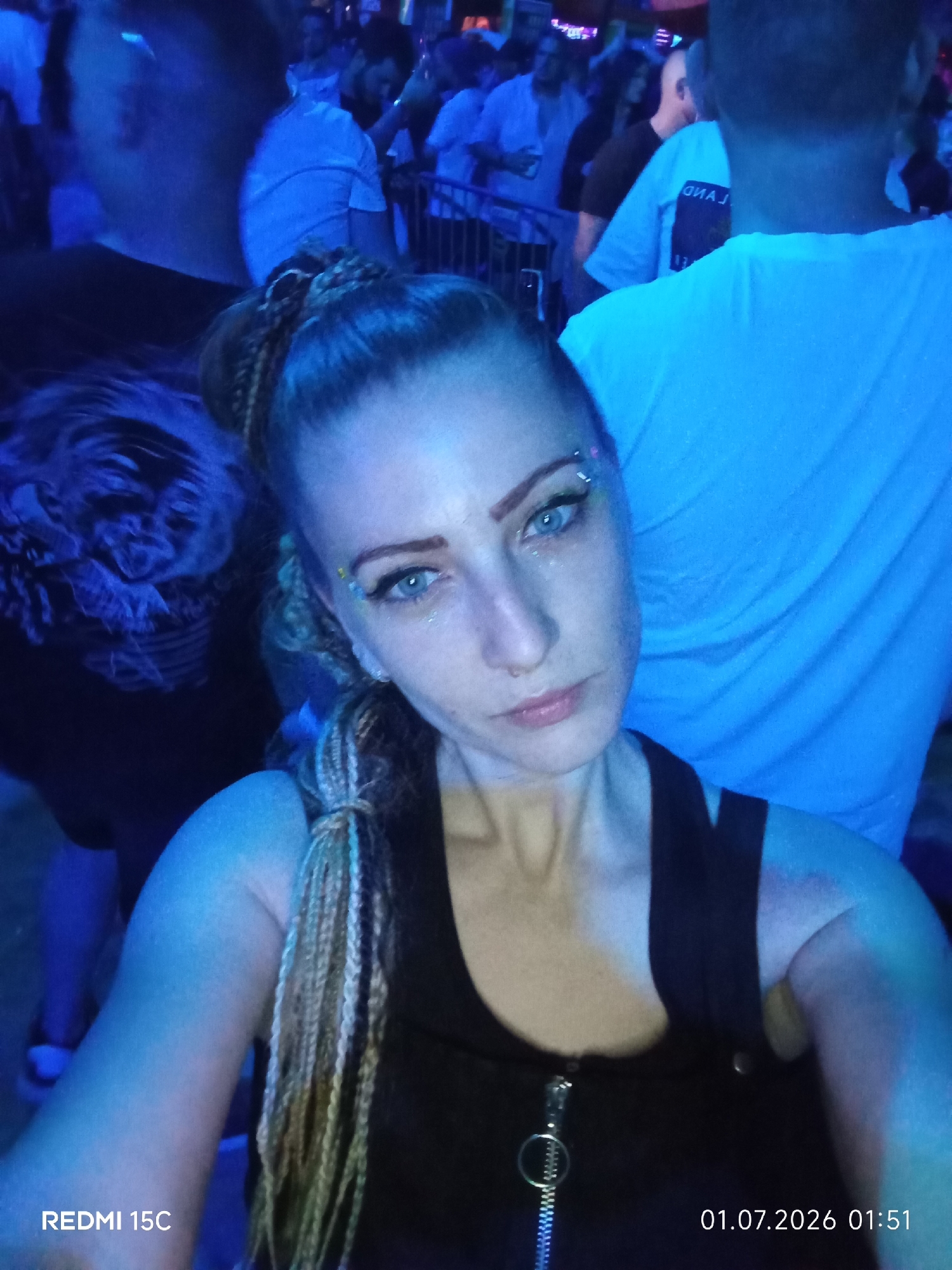 An outdoor, nighttime scene at an event, possibly a festival or concert, is captured in this selfie taken by a woman. She is in the foreground, looking towards the camera, with a slight smile. She has blonde braided hair and is wearing a black tank top. The background is filled with a crowd of people, some illuminated by blue and white lights. The overall atmosphere appears lively and energetic. There is a date and time stamp in the bottom right corner of the image, indicating "01.07.2026 01:51".