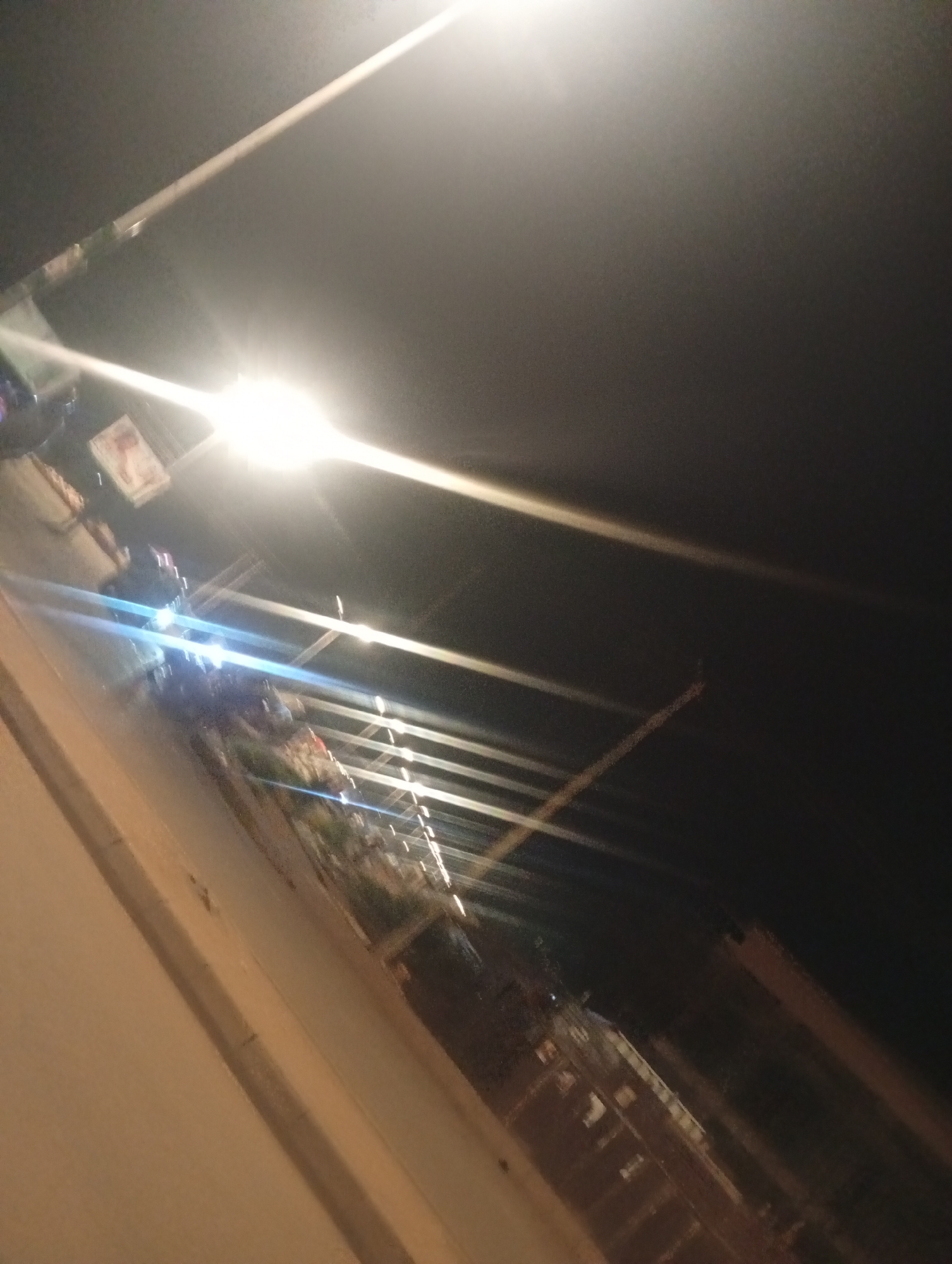 In the city of Bukuru, Nigeria, at night, an angled shot captures a street scene dominated by bright artificial lights. A person on a motorcycle, illuminated by headlights, is visible. The background shows a low-rise structure, possibly a building with multiple levels, and railings line the walkway. The overall ambiance is one of urban nighttime activity.