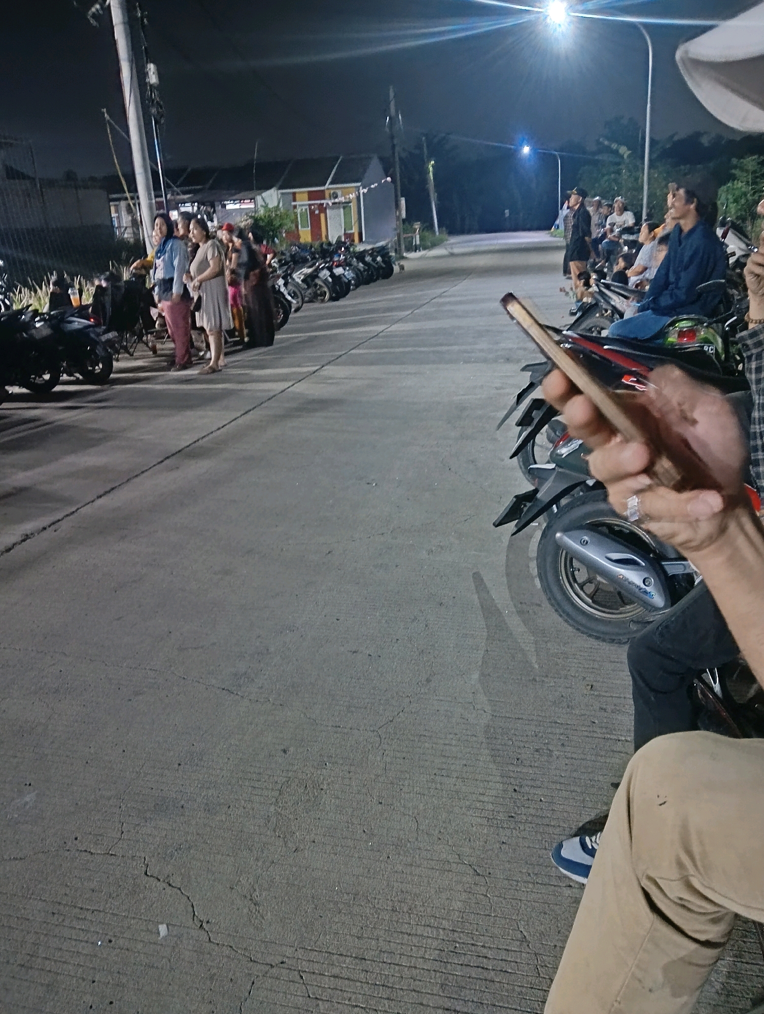 A group of people are standing and sitting next to parked motorcycles on a paved road at night in Cikasungka, Indonesia. The scene is illuminated by artificial lights, including a bright streetlamp in the background. Several individuals are standing together, some appear to be in conversation, while others are seated on motorcycles. A hand holding a smartphone is prominently featured in the foreground. The environment suggests an outdoor gathering or event, with buildings visible in the distance.