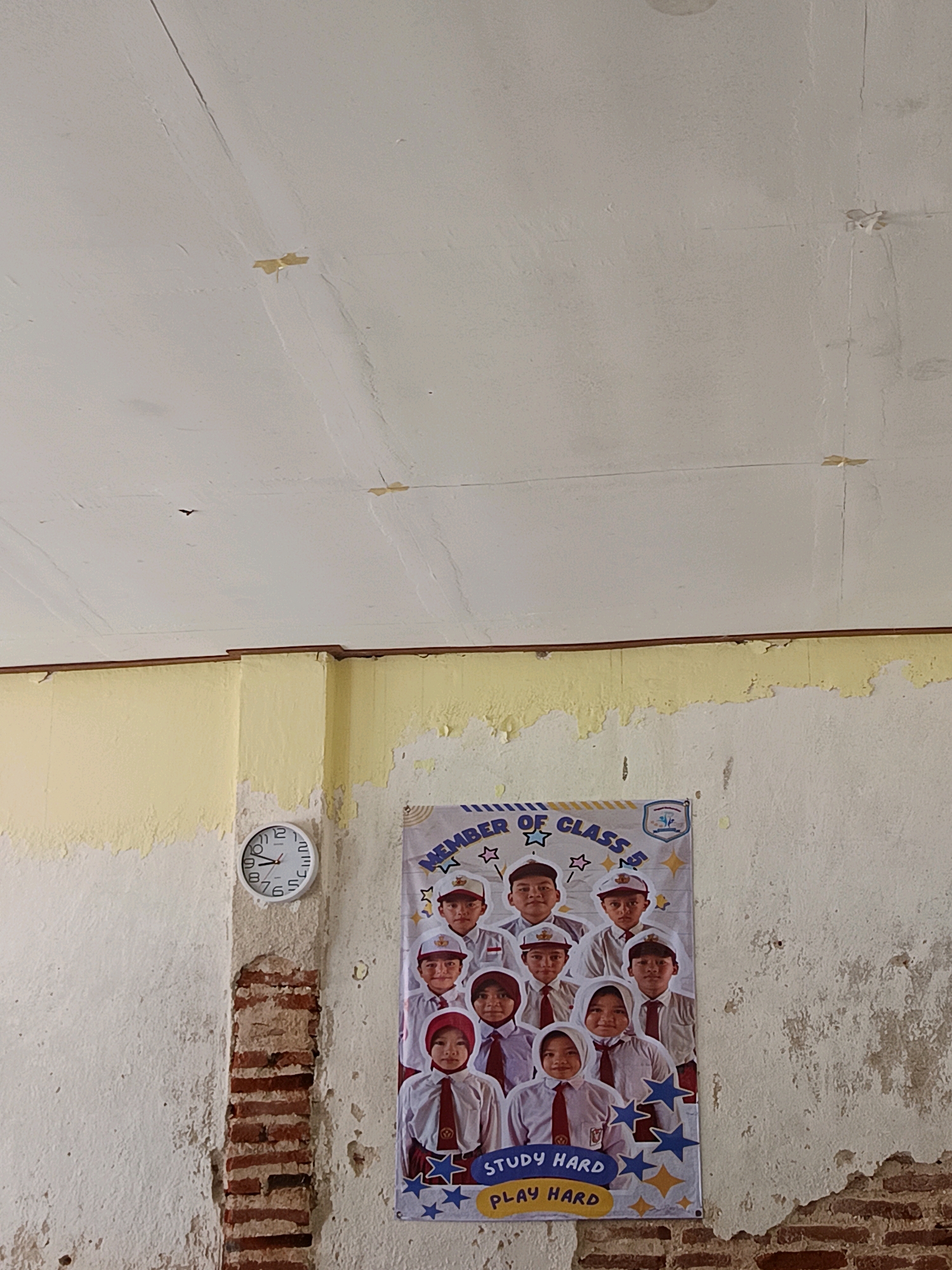The image depicts a classroom or hallway setting, likely in Labuhan, Indonesia. A poster of a class photo, featuring students in uniform, is displayed on a weathered wall. The poster has the words "MEMBER OF CLASS 5" and "STUDY HARD PLAY HARD" printed on it, along with decorative stars. To the left of the poster, a white analog clock is mounted on a textured, off-white wall. The wall above the clock and poster is painted a pale yellow and shows signs of peeling and water damage. The ceiling is a pale white with visible cracks and some yellowed tape. The overall environment appears aged and possibly neglected. There are no individuals present in the scene other than those in the poster.