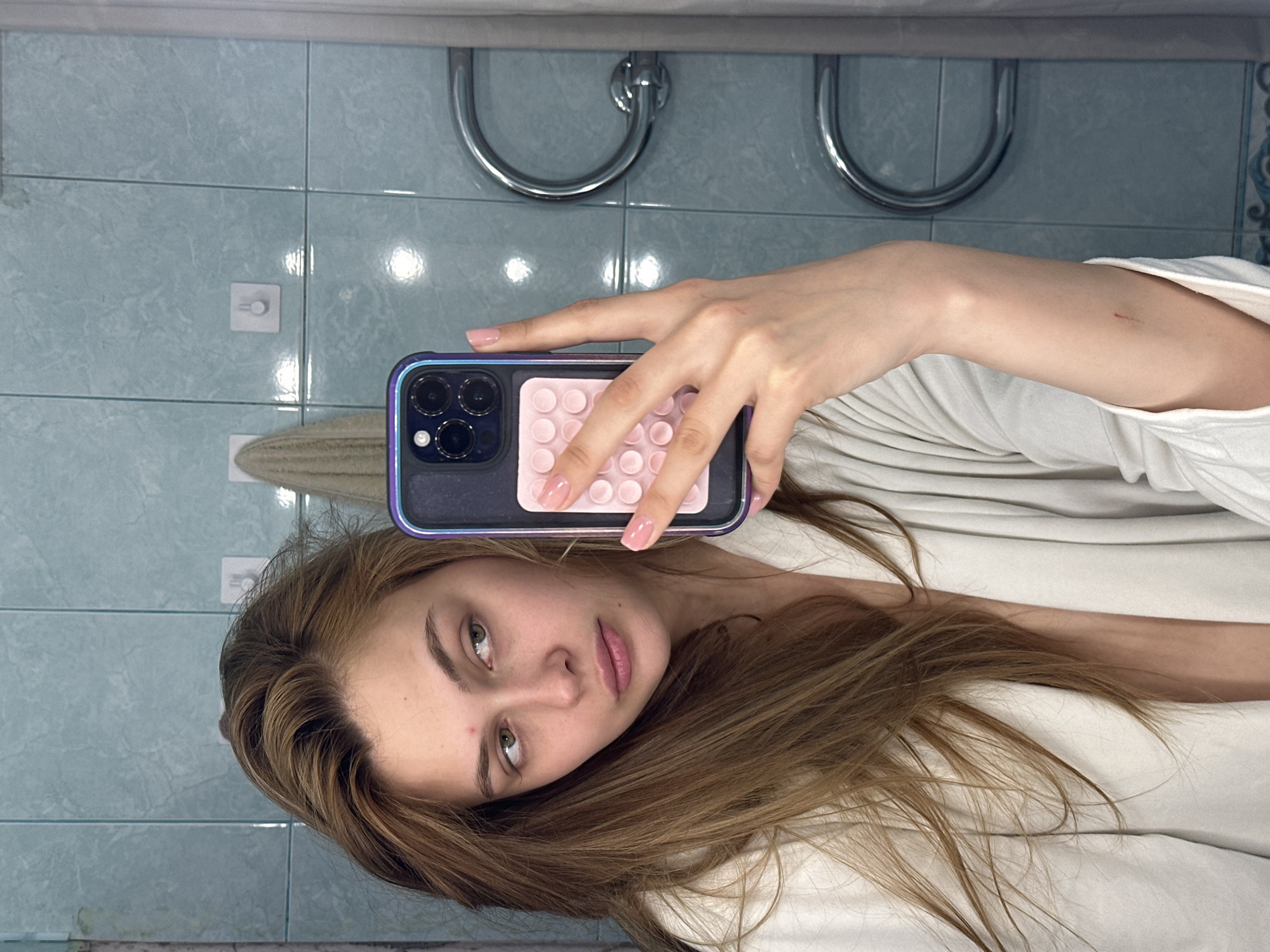 A young woman with long brown hair takes a selfie in a bathroom. She is wearing a white shirt and holding a smartphone with a pink case. The background shows a tiled wall, a towel rack, and a towel. This information does not provide specific details to conclude the location is Krasnoyarsk, Russia.