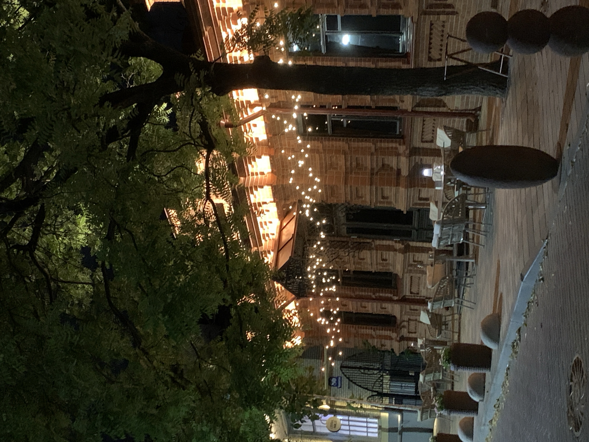 An outdoor seating area at night is illuminated by strings of warm-toned fairy lights draped across the facade of a brick building and a large tree with dense foliage in the foreground. The building, made of orange-red brick with decorative architectural elements, has large windows. Tables and chairs, appearing to be made of wicker or rattan, are arranged on a paved patio in front of the building, some with umbrellas. Several large, dark, oblong sculptures are placed on the patio. To the left, a sidewalk with scattered leaves leads away from the seating area. The scene is dark, with the primary light sources being the building's artificial lighting and the fairy lights.