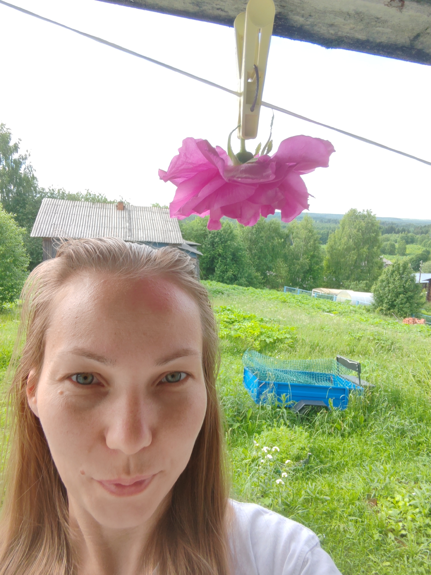 A woman with light brown hair and green eyes is taking a selfie in an outdoor setting. She has a red mark on her forehead. In the background, there is a rural landscape with a wooden house, trees, and a greenhouse. A pink rose is hanging from a clothesline with a clothespin above her head. A blue trailer with a green net is visible in the grassy field. The scene appears to be in a village in the Чухлэм district of Russia.