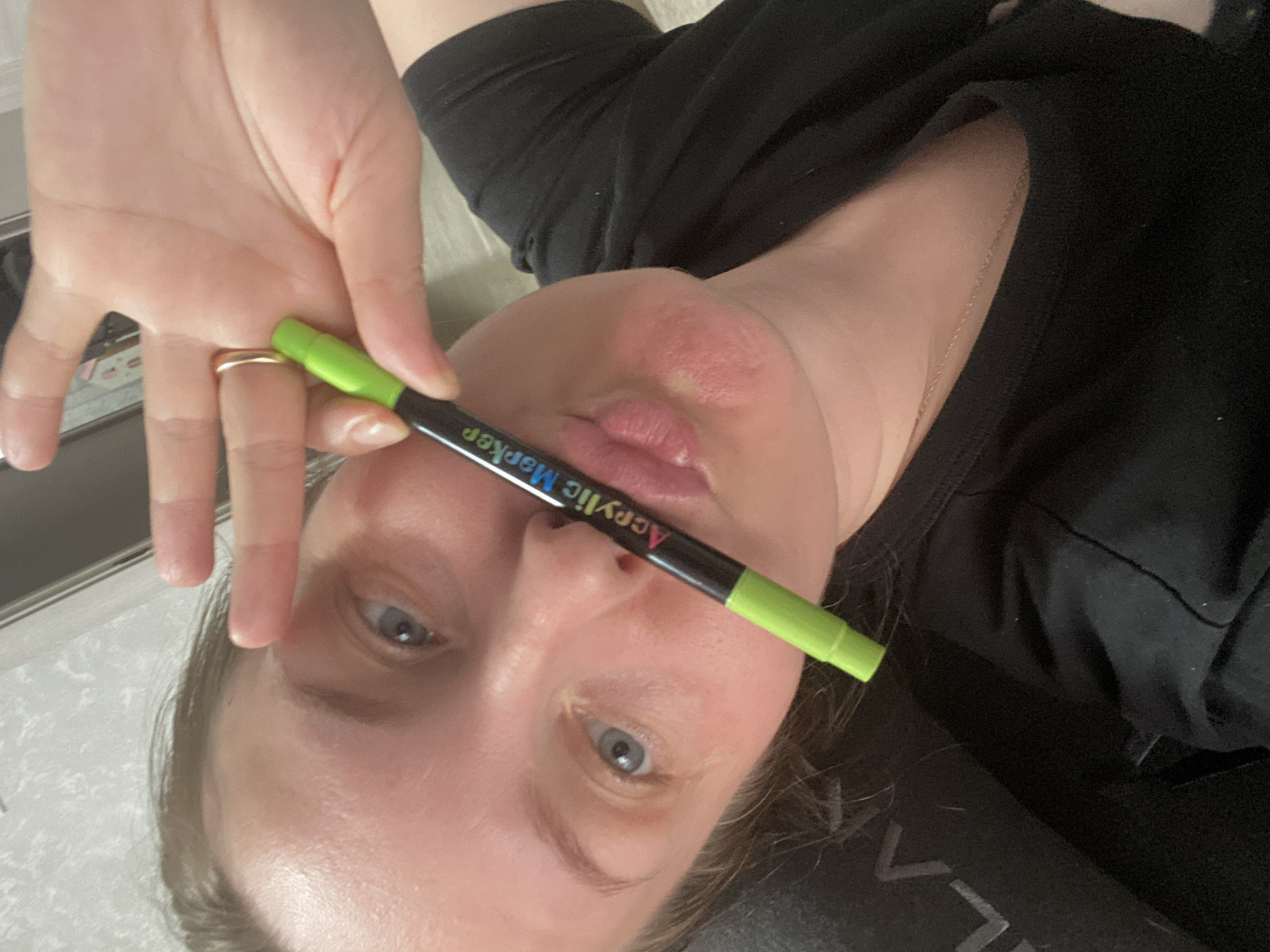 This media file is a close-up, rotated portrait of a person captured in a selfie-style, appearing to be lying down and looking towards the camera. The image is oriented sideways, with the person's head to the right.
The central subject is an adult with light skin and light blue-grey eyes. Their hair, visible around the face, appears to be a light brown color. They are wearing a black short-sleeved top. A thin, gold-colored chain necklace is visible on their neck, and a simple gold ring is worn on the ring finger of their left hand.
The person holds a green and black "Acrylic Marker" horizontally across their face with their left hand, positioned just above their mouth. The marker has a bright green cap and tip, with a black barrel. Clearly visible text on the marker barrel includes "Acrylic Marker" in white, red, and blue lettering. The person's lips are slightly pursed.
The activity depicted is a posed photograph. The background is softly blurred, suggesting an indoor setting with neutral-colored surfaces. No specific time of day or outdoor weather can be determined. The person's expression is neutral, possibly playful due to the slight pucker of the lips. The location context provided for this media is Noviy, Russia.