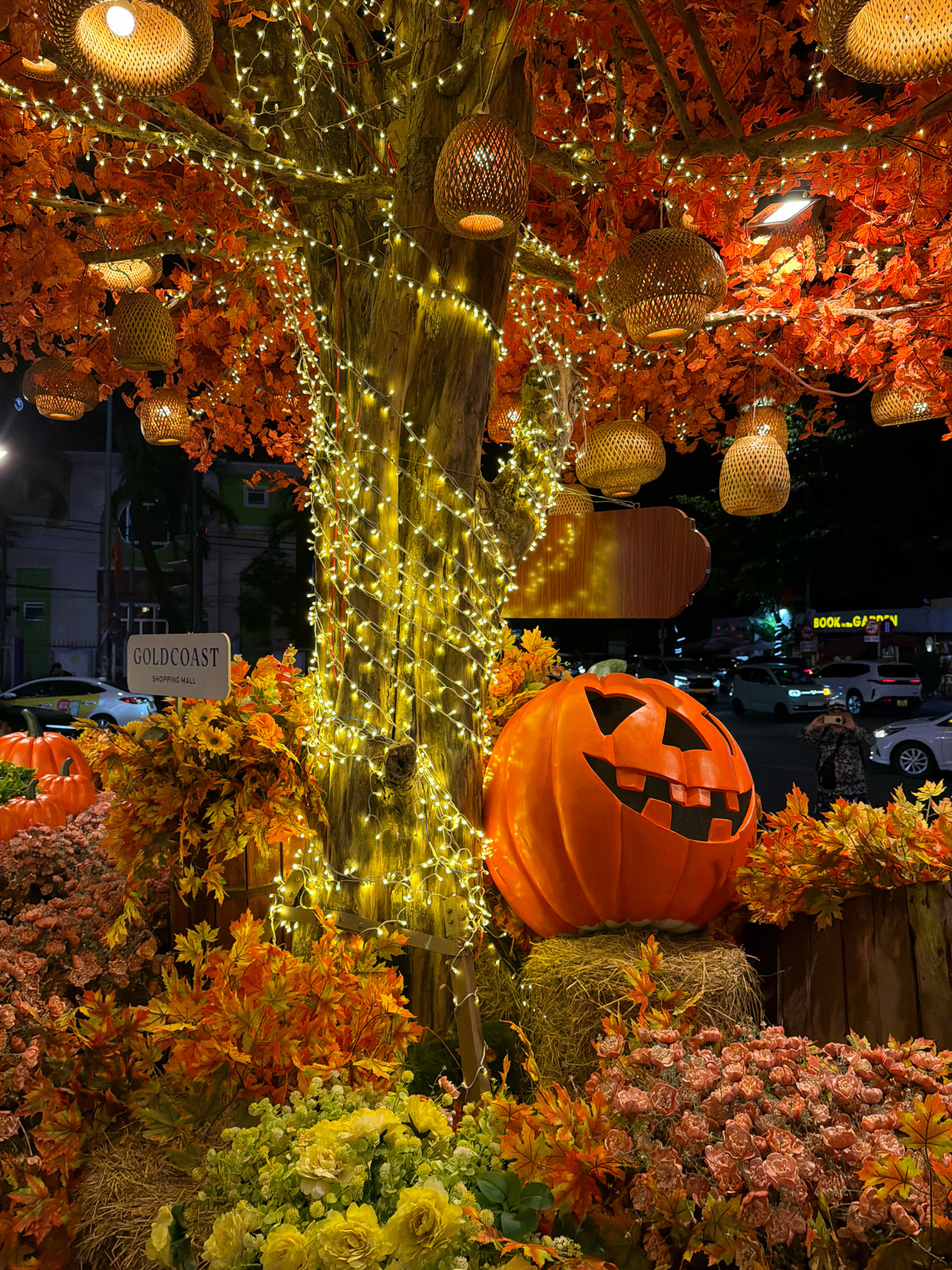 This image depicts a festive Halloween decoration in front of the Goldcoast Shopping Mall in Nha Trang, Vietnam. The primary focus is a large, carved pumpkin with a menacing grin and empty eye sockets, placed on a bale of hay. Behind the pumpkin, a tree is adorned with numerous strands of warm yellow string lights and several woven hanging lanterns. The tree is also decorated with artificial orange and red maple leaves, creating a vibrant autumnal atmosphere. Surrounding the base of the tree and pumpkin are dense arrangements of artificial flowers in shades of pink, yellow, and orange, interspersed with more artificial leaves. In the background, the street is visible with cars and a few pedestrians, and signs for "Goldcoast Shopping Mall" and "BOOK .n. GARDEN" are illuminated. The scene is set at night, with the artificial lighting creating a warm and inviting glow.