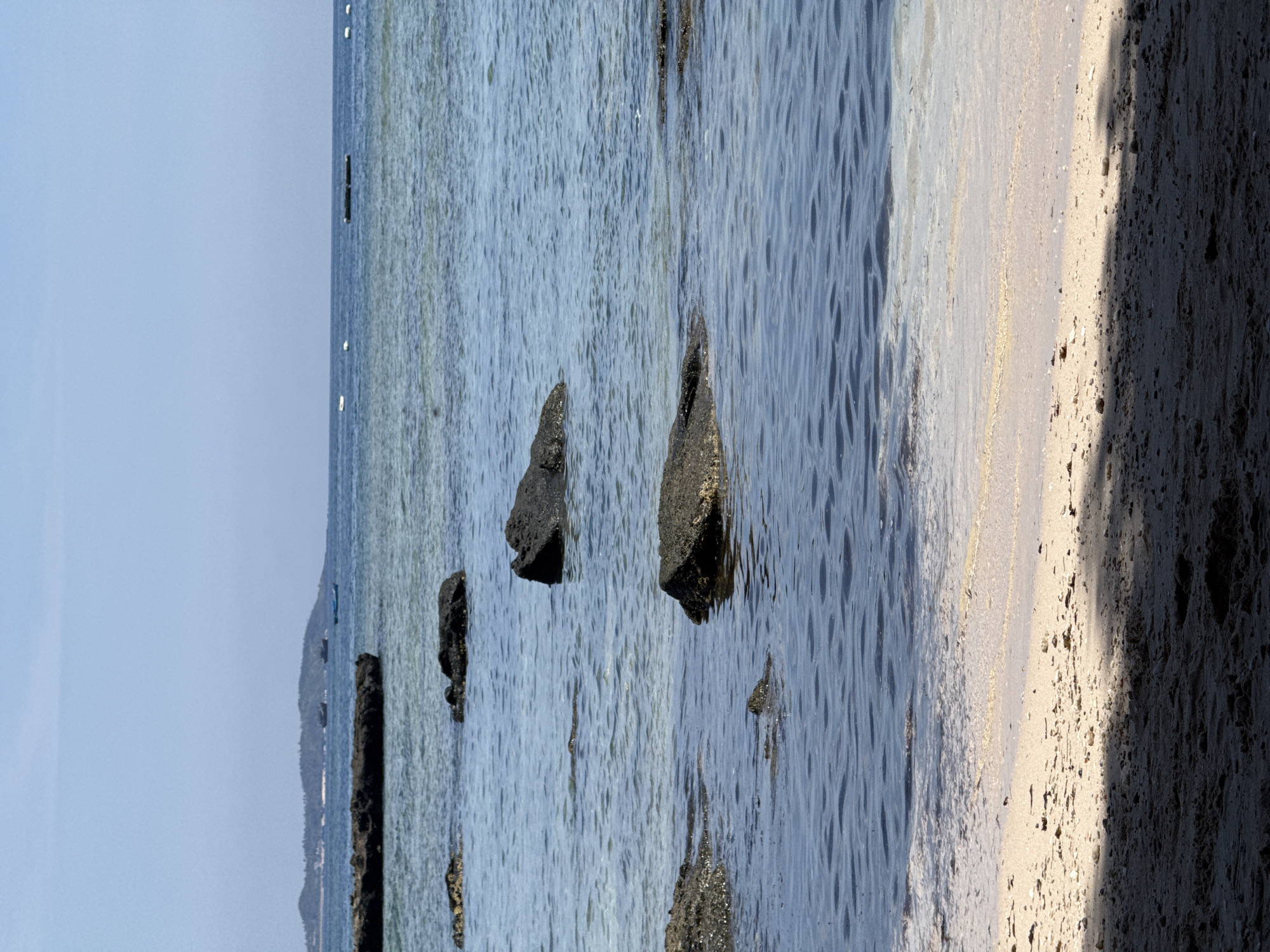 The image depicts a serene coastal scene on Lý Sơn Island, within the Lý Sơn Special Administrative Region of Vietnam. The foreground is occupied by a sandy beach, partially covered in shadows, with scattered pebbles and debris. The sea gently laps at the shore, creating a shallow, shimmering waterline. Several dark, rugged rocks emerge from the calm, blue water at varying depths. In the middle ground, more rocks are visible, some partially submerged. The horizon is marked by the distant outline of a lush, green island or peninsula, dotted with trees and a few small boats. The sky above is a clear, pale blue, with a few faint white clouds near the horizon. The overall impression is one of tranquility and natural beauty.