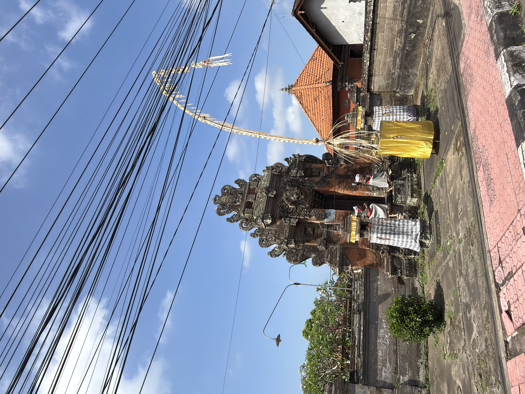 In Blahbatuh, Indonesia, a traditional Balinese temple gate stands prominently. The gate, constructed from weathered stone and adorned with intricate carvings, is a focal point of the scene. Two stone pillars flank the entrance, each draped in a checkered cloth. To the right of the gate, a tall, arched decoration, likely part of a religious offering or celebration, reaches upwards, adorned with yellow fabric and woven palm fronds. A smaller decorative element, also made of palm fronds, is visible at the base of this structure. In front of the temple, a small, dense green bush grows in a patch of cracked pavement. Behind the gate, a red-tiled roof of a nearby building is visible, along with a stone wall and lush greenery. Overhead, a network of black electrical wires crisscrosses the bright blue sky, which is dotted with scattered white clouds. The foreground features a paved walkway with a distinctive red and white striped curb.
